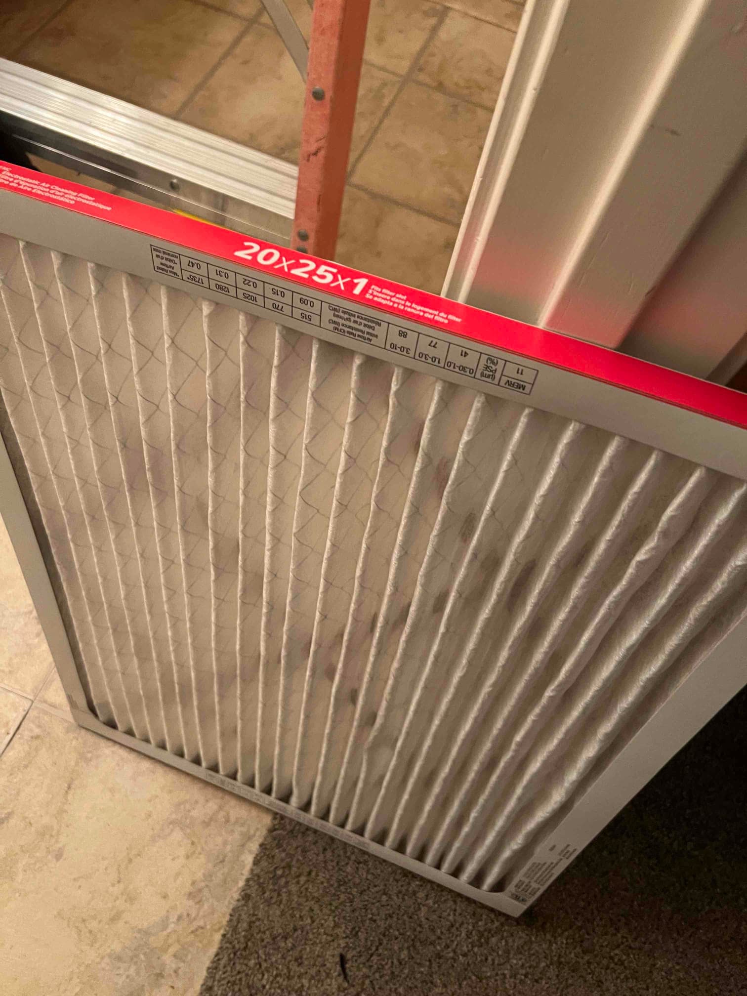 Pleated air filter labeled 20x25x1 resting near a return grille frame indoors.