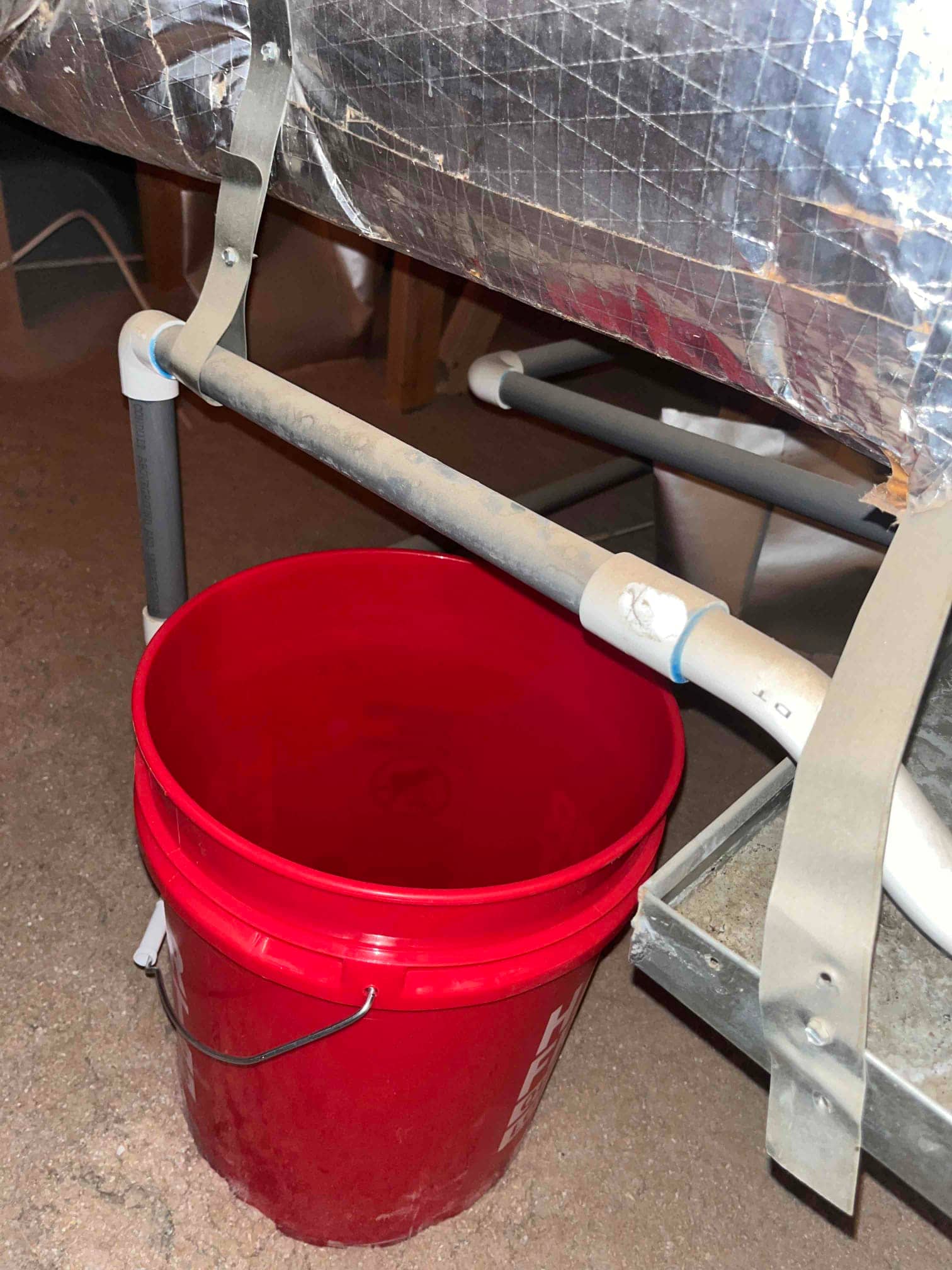 PVC drain line discharges above a red bucket beneath insulated duct and support frame.