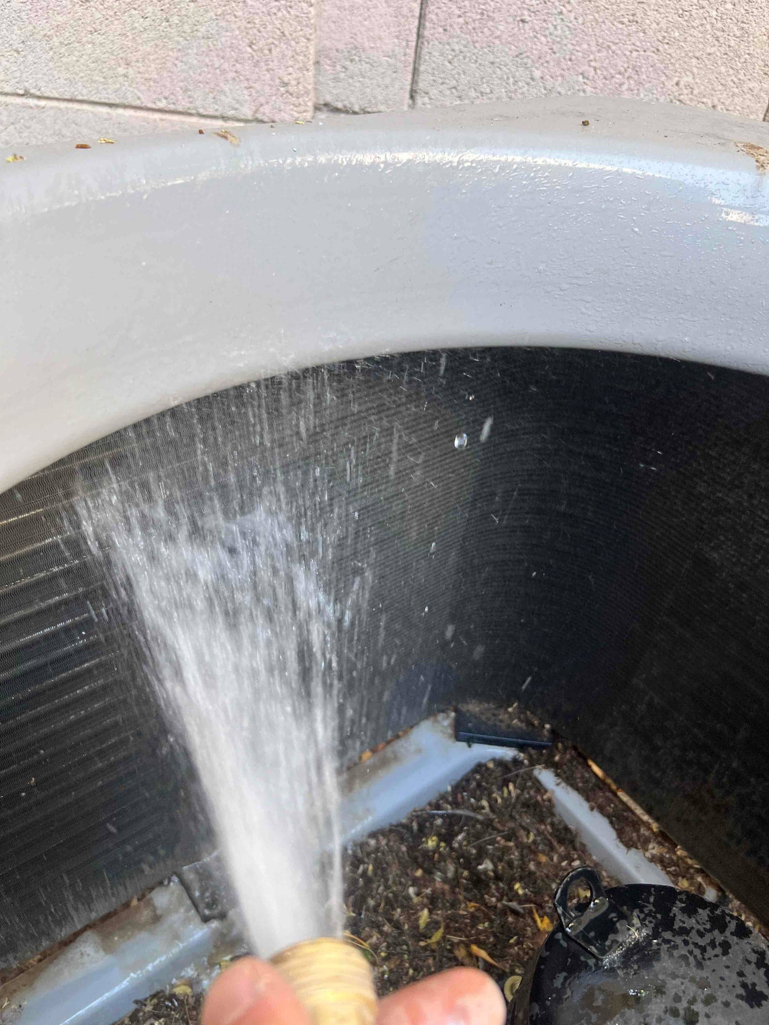 Water hose sprays the inside of an outdoor condenser coil, with accumulated debris in the base pan.