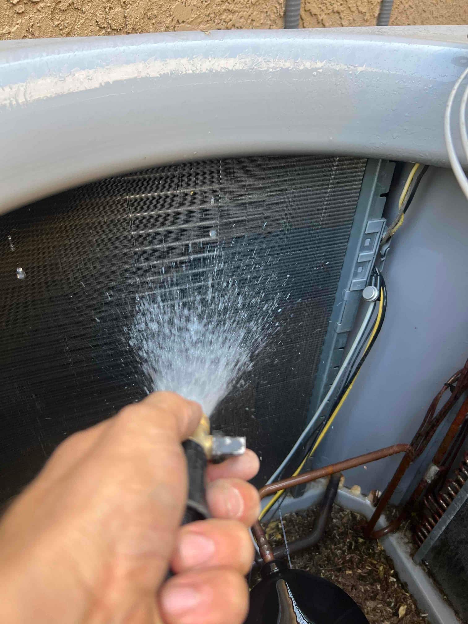 Water spray from a hose directed onto the outdoor condenser coil with wiring and copper refrigerant lines visible.