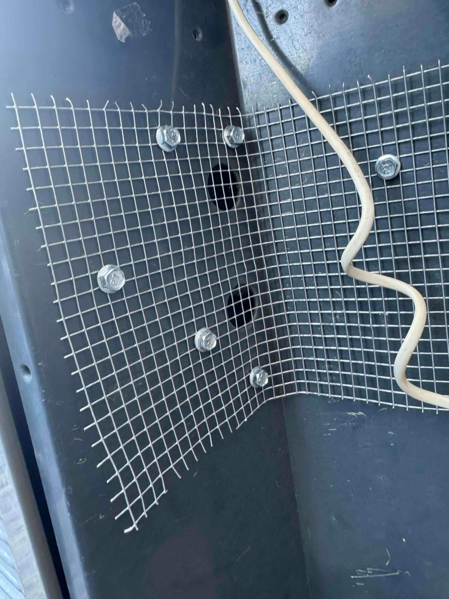 Wire mesh screen fastened with bolts inside equipment cabinet, with a beige wire routed alongside.