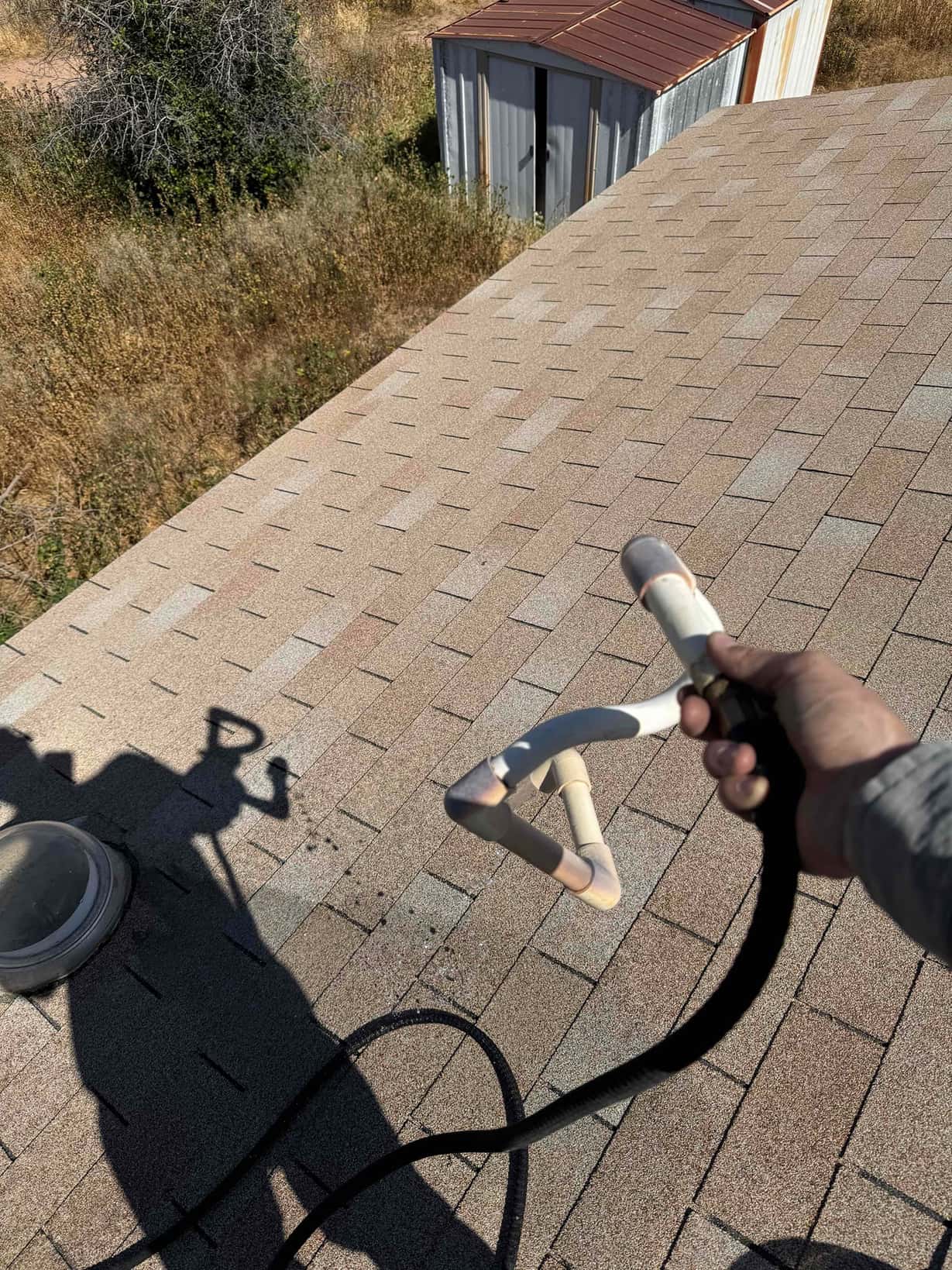 Hand holds a spray wand with hose on a shingle roof beside a small skylight.