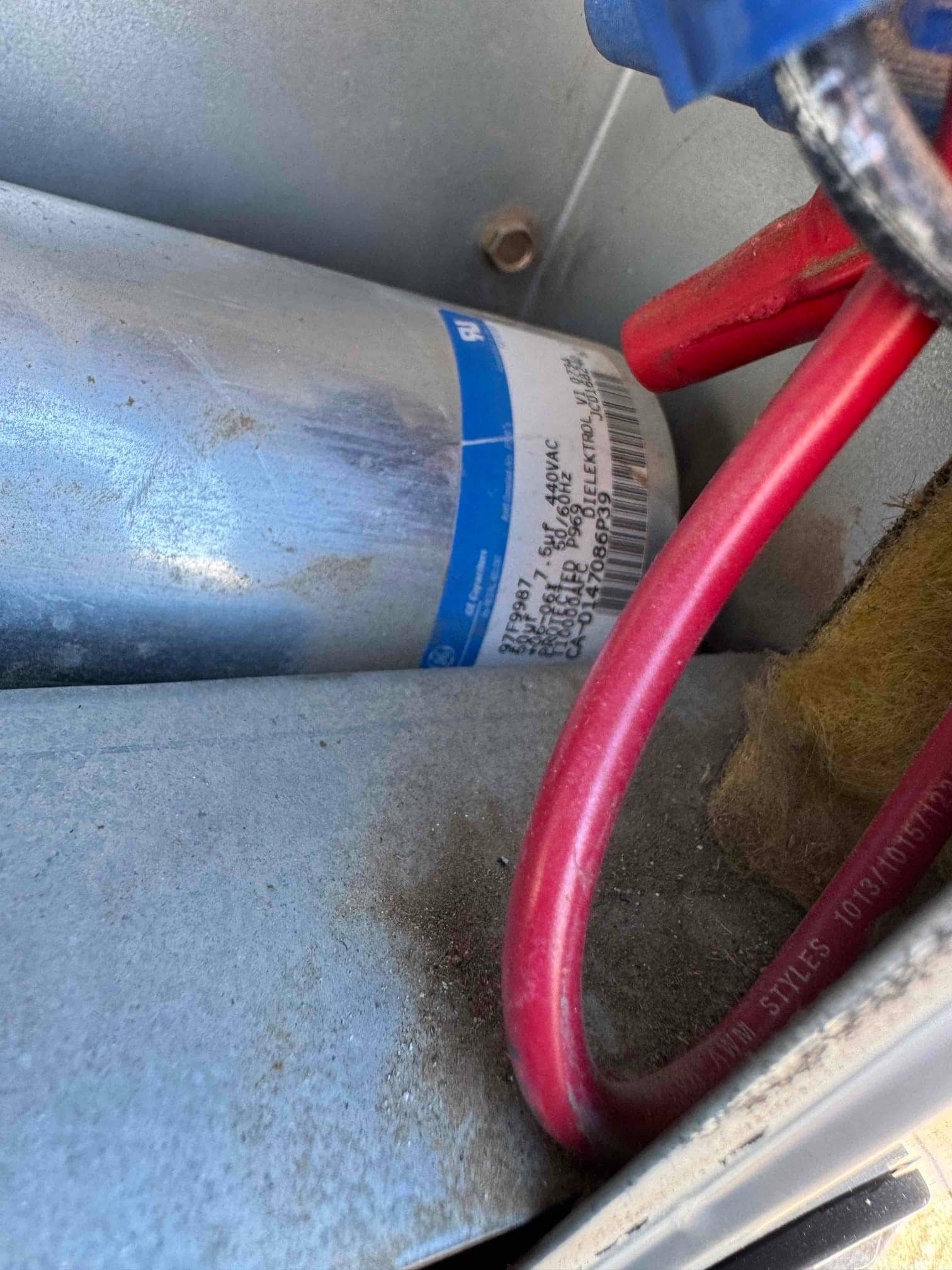 Metal can run capacitor with red wiring connected inside the outdoor unit cabinet, with visible dust on surrounding surfaces.