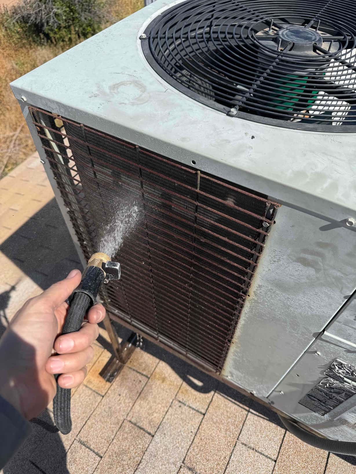 Outdoor condenser coil on rooftop unit is rinsed with a hose; fan guard and coil fins visible.