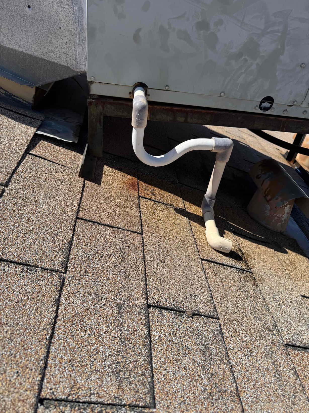 PVC condensate drain assembly exiting a rooftop HVAC unit over shingle roof, with elbows and visible rust staining below.