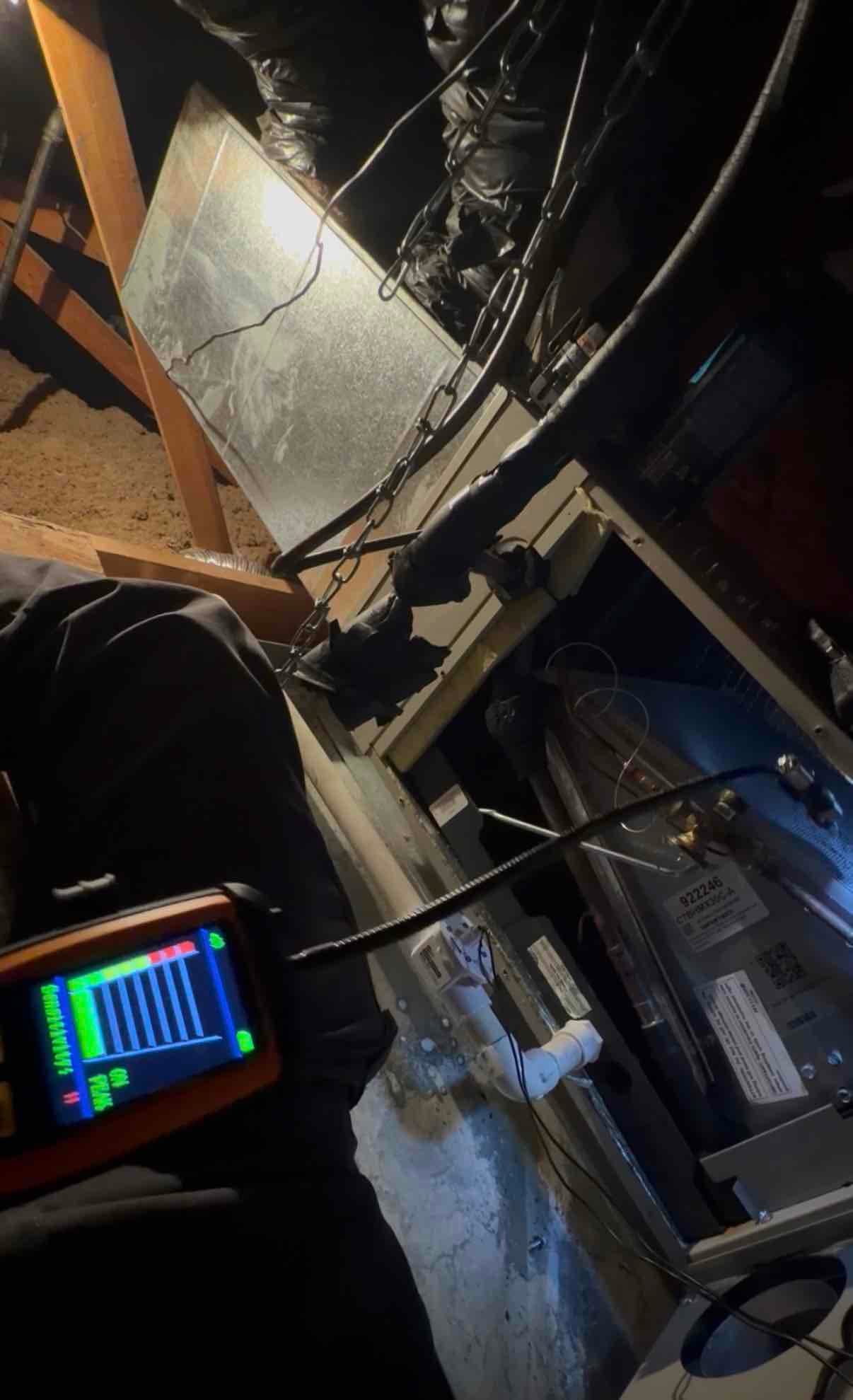 Air handler cabinet open in attic with insulated refrigerant lines and PVC condensate drain; technician uses a handheld probe meter near the coil.