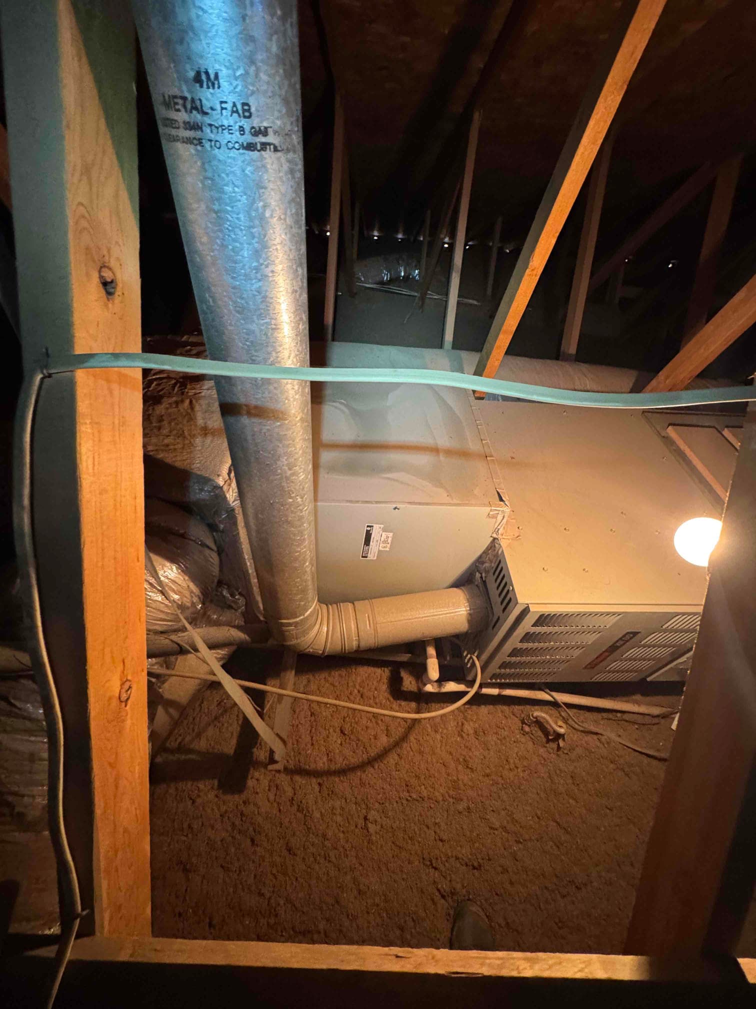 Attic horizontal furnace with vent piping, flexible duct connections, and service light illuminating surrounding framing.