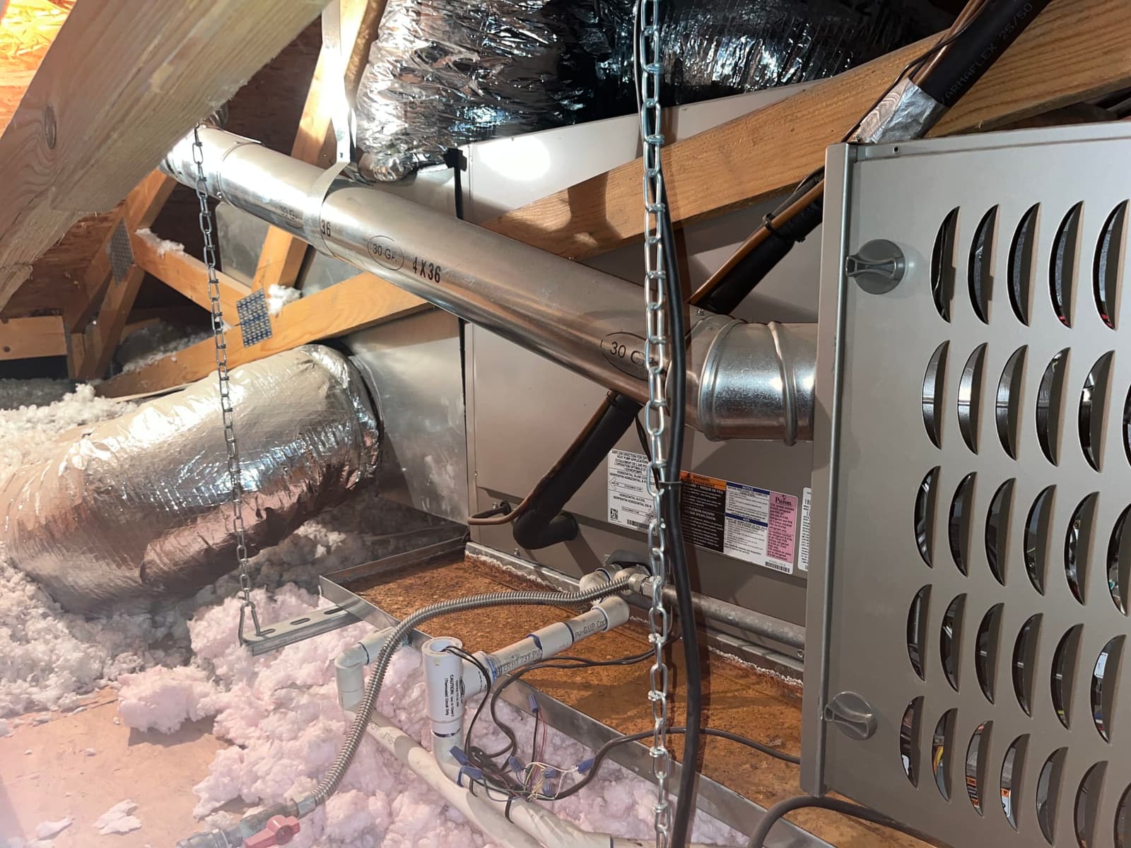Attic-installed furnace with metal vent pipe, insulated flex duct connections, condensate drain trap, service disconnect, and surrounding blown-in insulation visible.