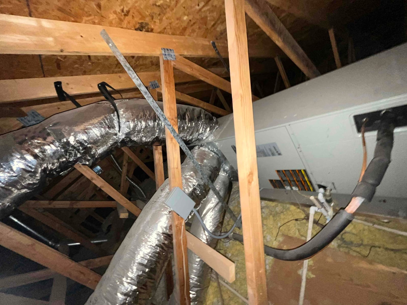 Attic-mounted air handler with insulated flex ducts, refrigerant lines, and condensate drain tubing visible among roof trusses.