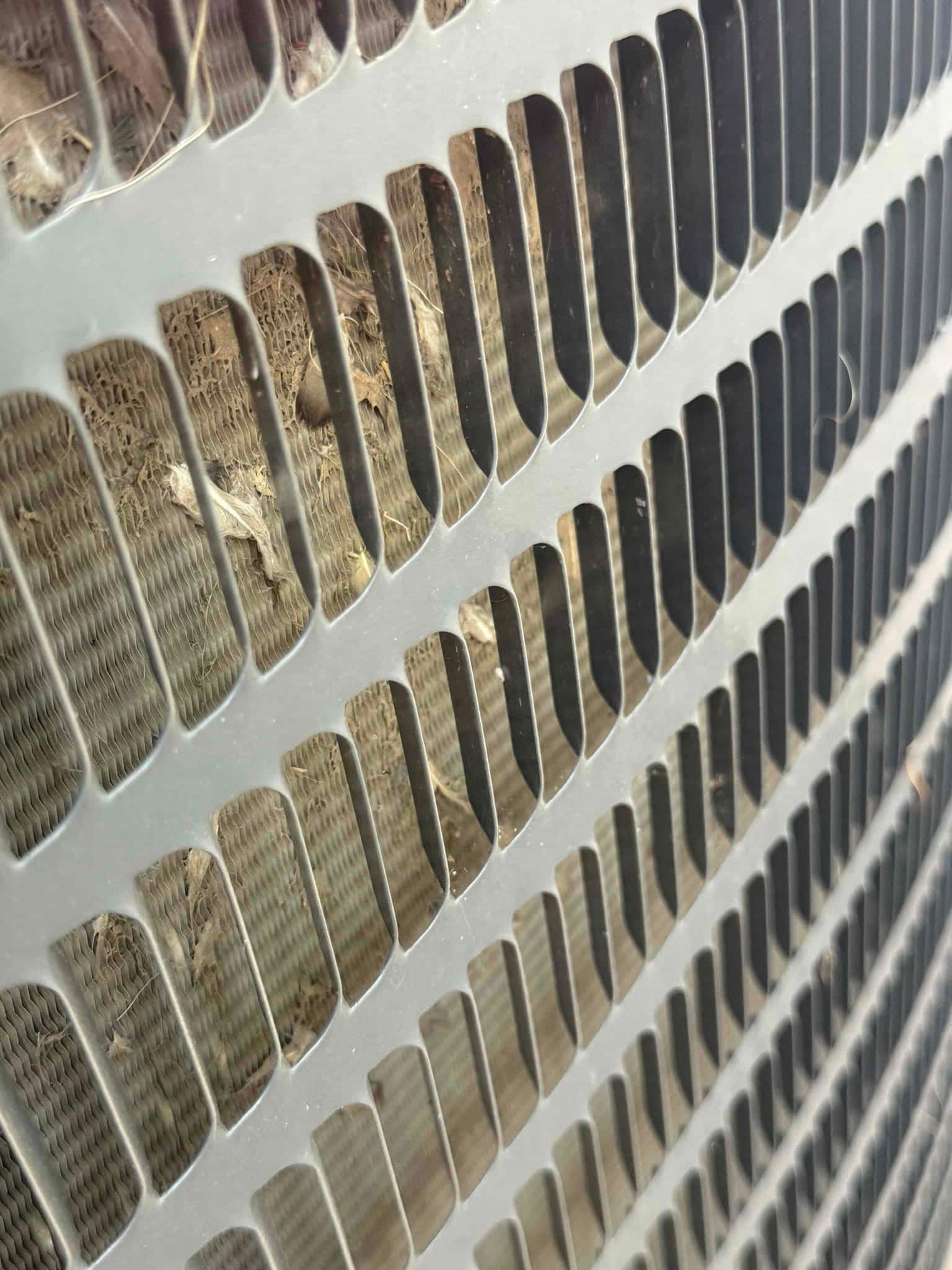 Close-up of an outdoor condenser’s metal grille showing coil fins behind it with leaves and debris.
