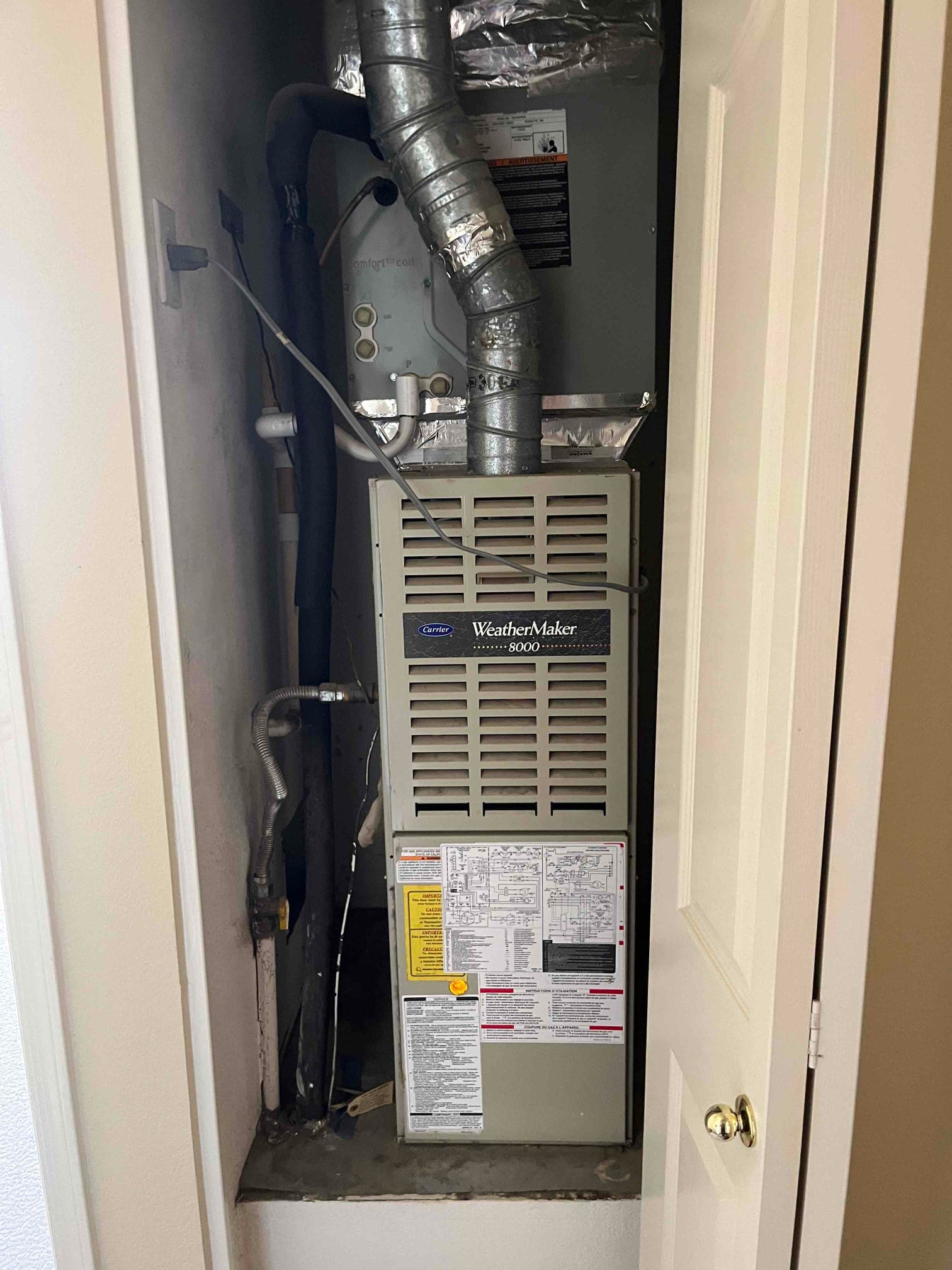 Closet-installed forced-air furnace with metal flue vent, evaporator coil cabinet, insulated refrigerant line, condensate drain, and service labels on the door.
