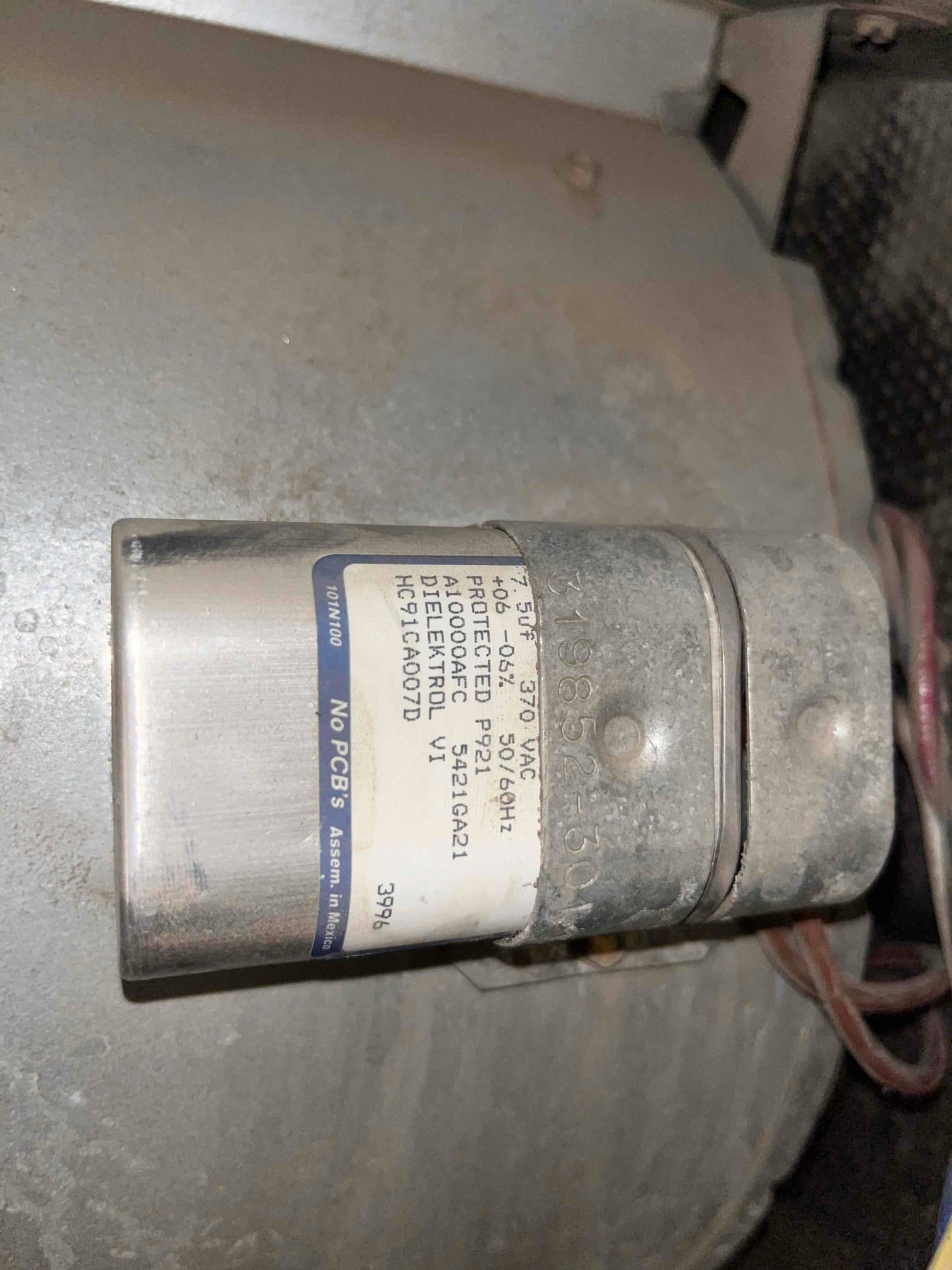 Cylindrical metal run capacitor mounted on the blower housing, with connected wires and visible dust on the surface.
