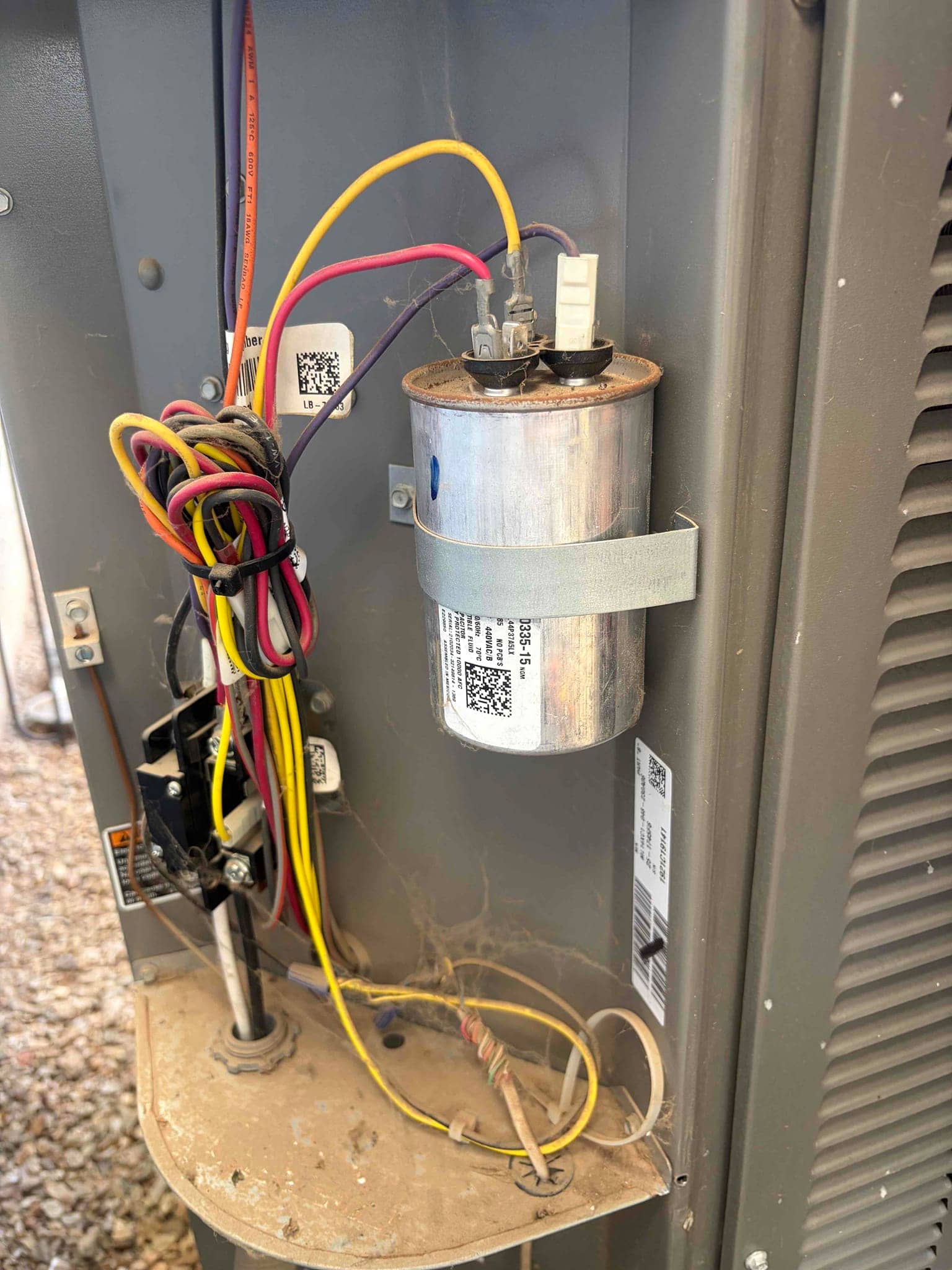 Cylindrical run capacitor mounted in the outdoor condenser panel with connected wires, nearby contactor, and visible dust and cobwebs.