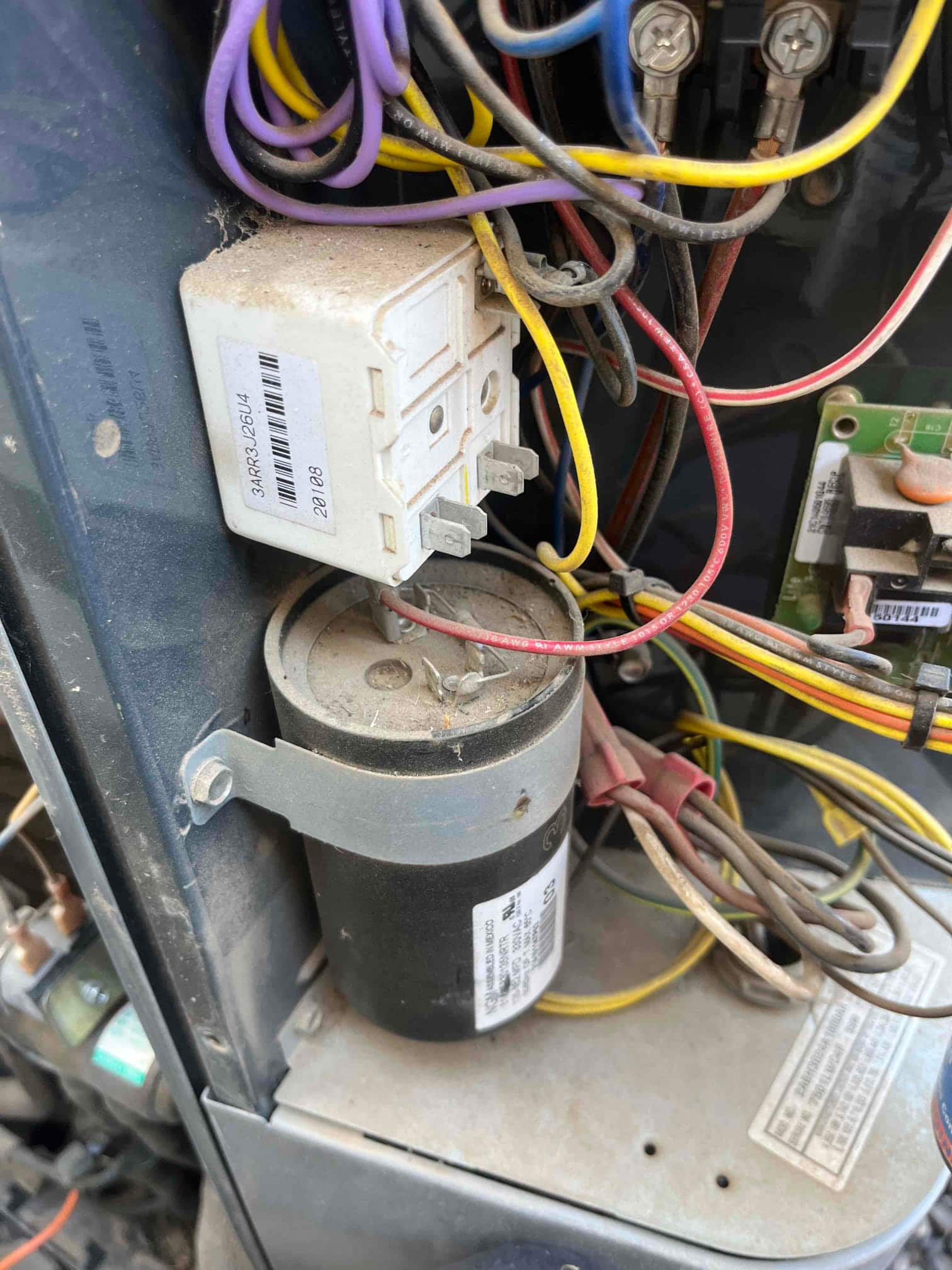 Cylindrical run capacitor mounted with strap, connected wiring and nearby relay inside condenser control compartment.