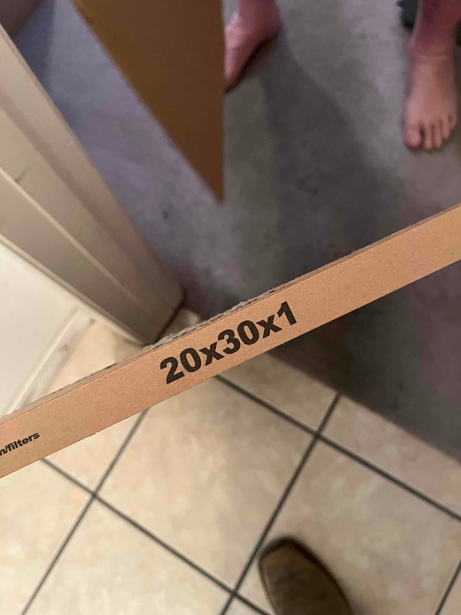 Disposable HVAC air filter edge showing size marking 20x30x1, held indoors near tiled floor.