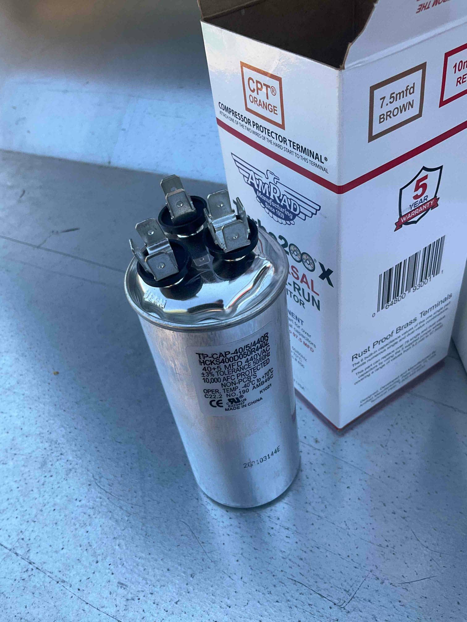 Dual-run capacitor with three top terminals, photographed next to its box on the outdoor unit panel.