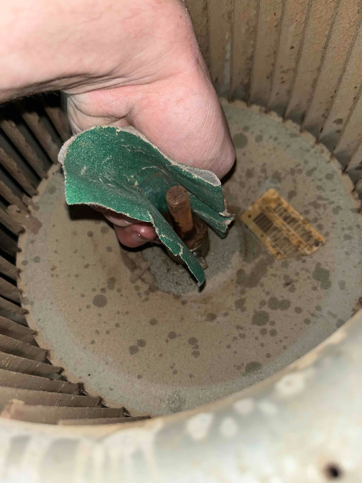 Dust-covered blower wheel interior with hand holding green abrasive pad near motor shaft.