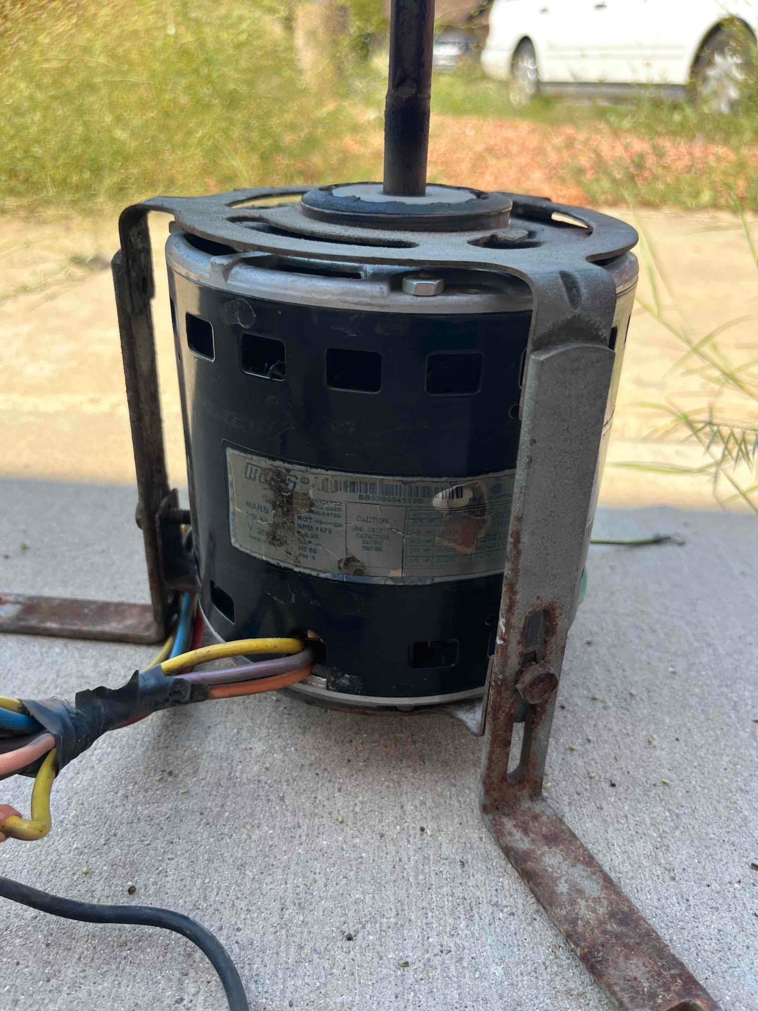 Electric fan motor on mounting bracket with rusted hardware and taped wiring, identification label visible on the housing.