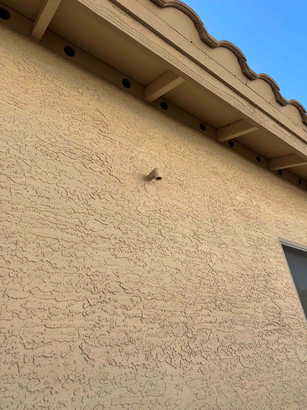 Exterior stucco wall showing a small angled drain outlet beneath the eave, near a window frame.