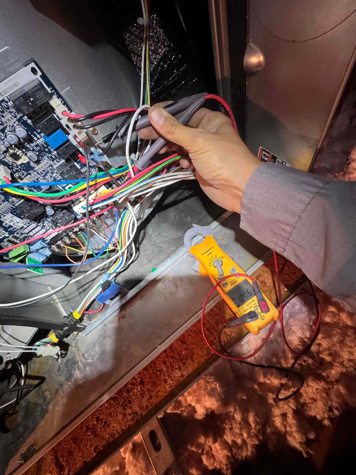 Furnace control board with multiple wires exposed while a technician holds test probes on the wiring harness.