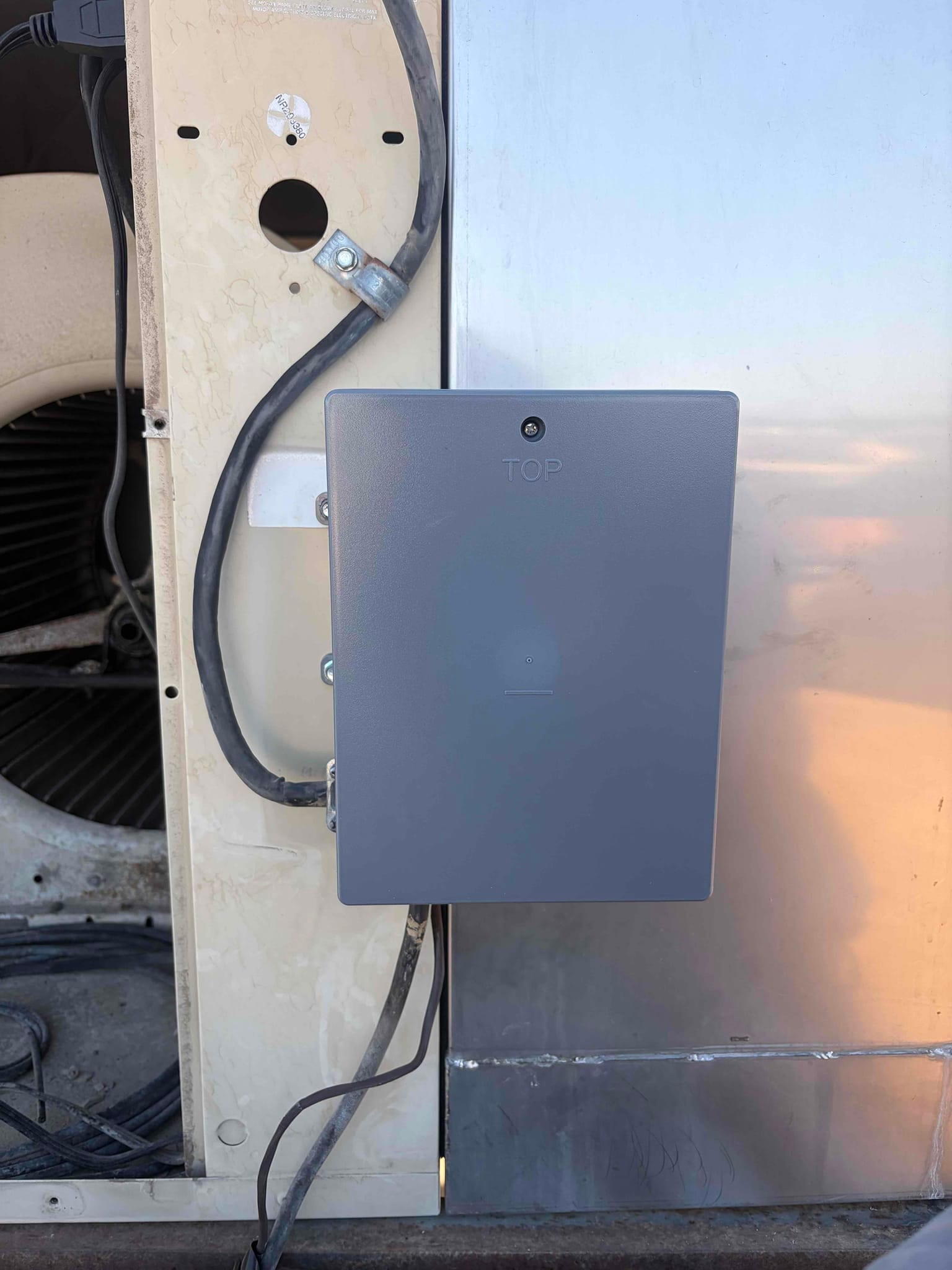 Gray electrical enclosure mounted with conduit and cables beside outdoor condenser housing and fan opening.