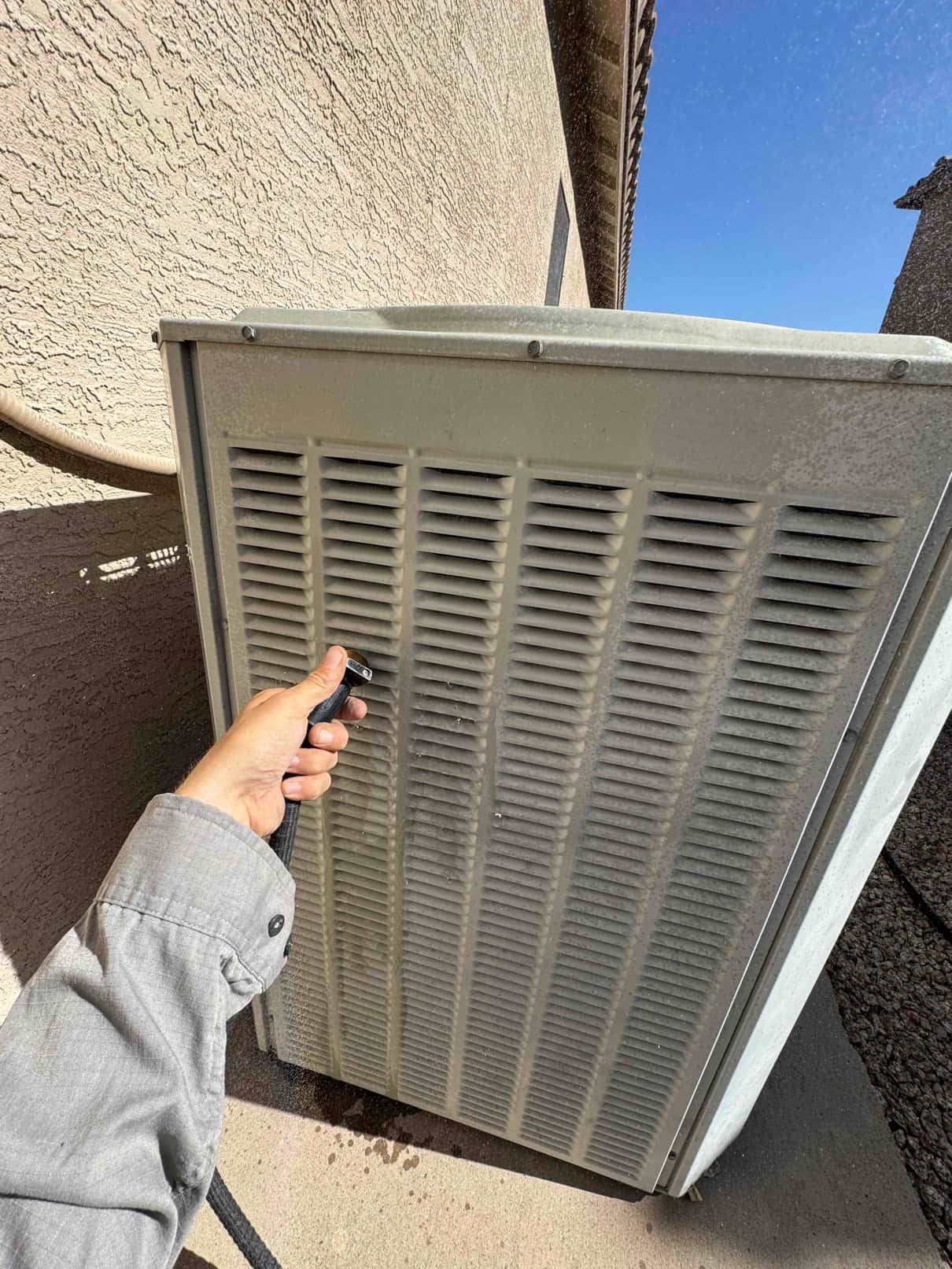 Hand uses a hose to rinse debris from the louvered side of an outdoor condenser unit.