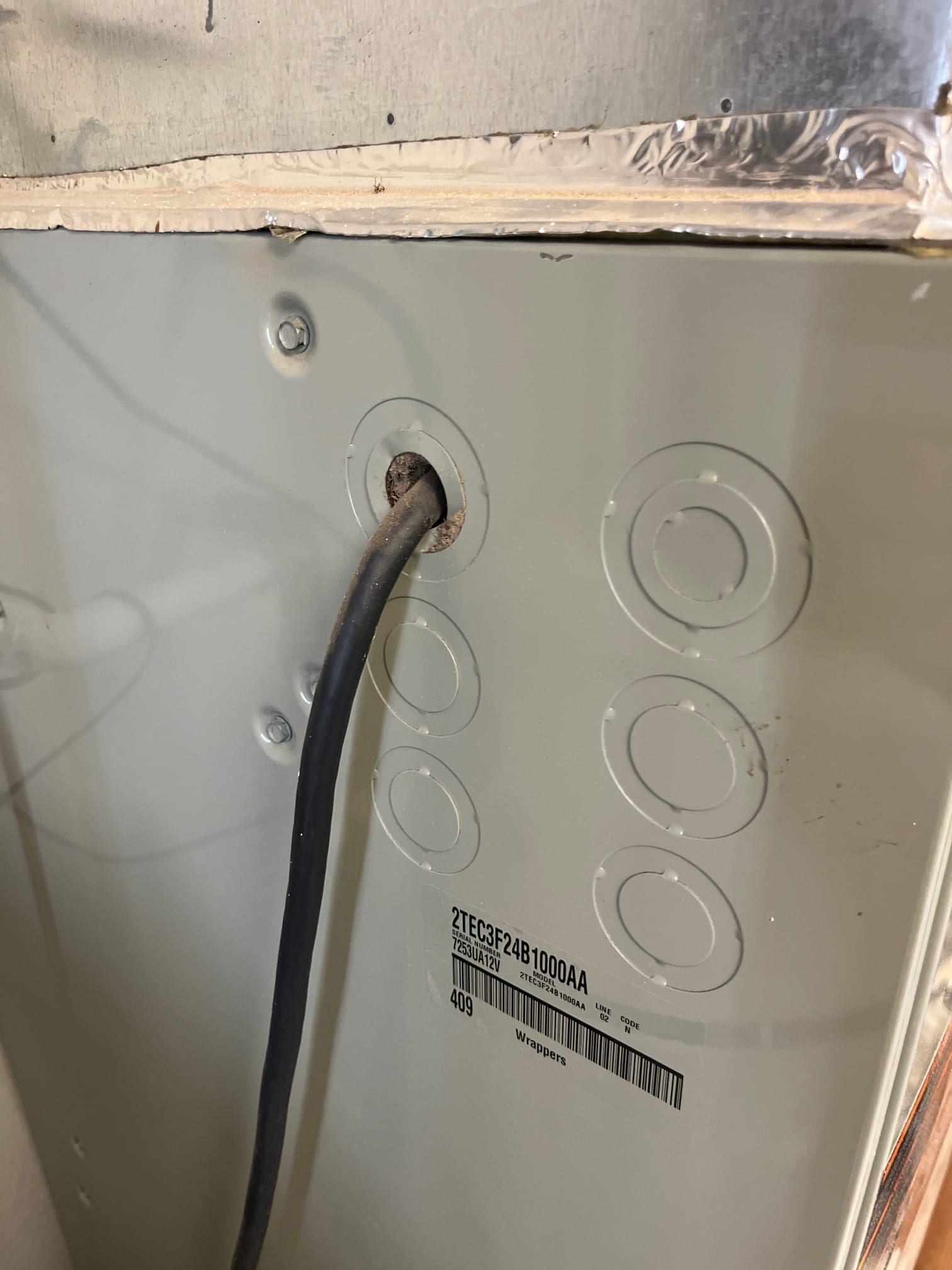 HVAC cabinet side panel with concentric knockouts and an electrical cable routed through one opening, foil-taped duct transition above.