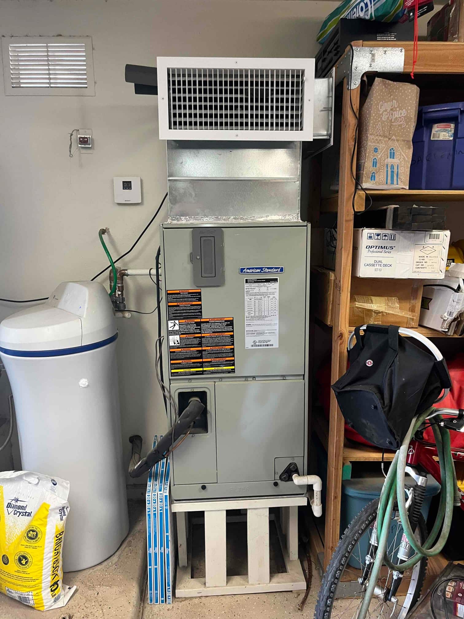 Indoor air handler on a stand with return grille above, insulated refrigerant line, and PVC condensate drain.