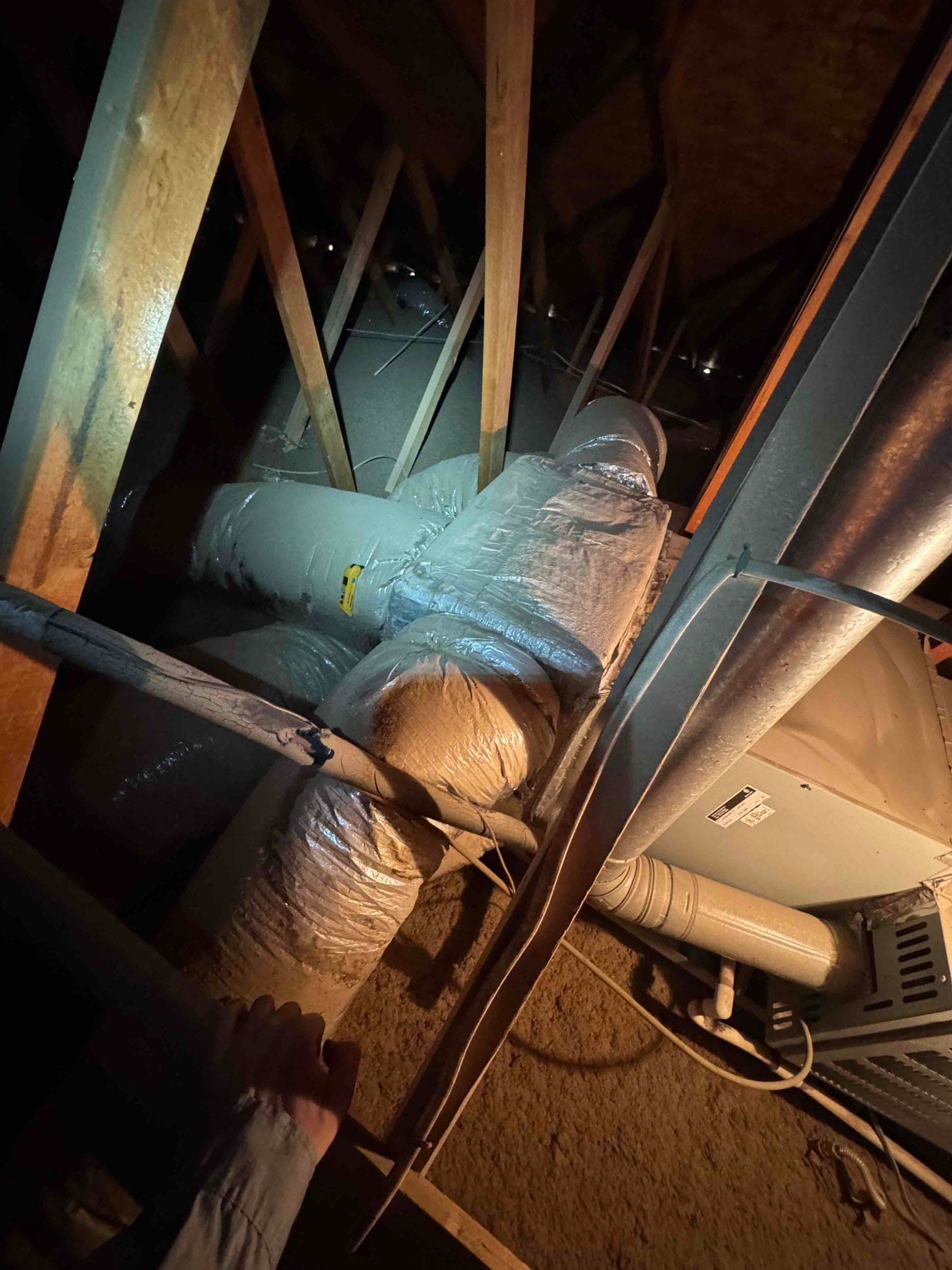 Insulated flexible ductwork connects to an attic HVAC unit, with drain piping and surrounding trusses visible in the space.