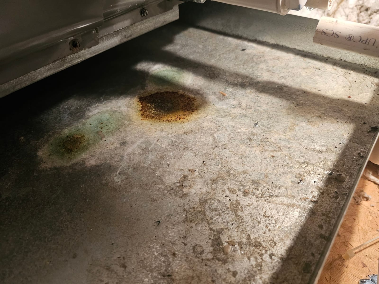 Metal drain pan under air handler with rust-colored and green discoloration and moisture marks.