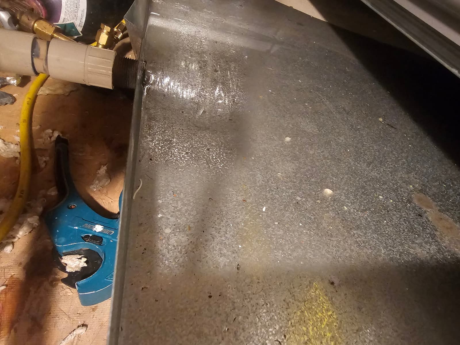 Metal drain pan with PVC condensate drain connection and visible water on the pan surface.