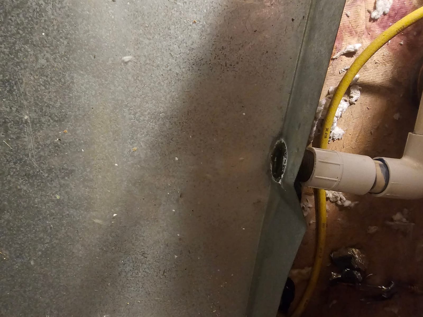 Metal HVAC cabinet side with PVC condensate drain connection and a yellow flexible line against the wall.