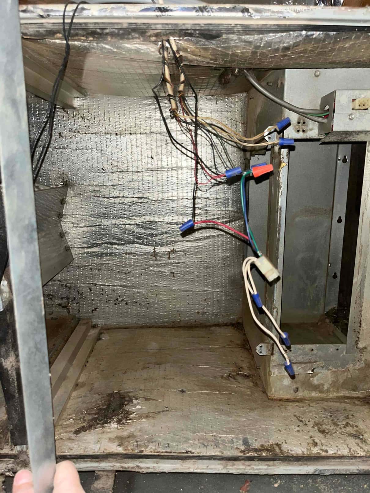 Open air handler cabinet interior showing loose electrical wiring with wire nuts, foil-faced insulation, and dirt and rust on the base.