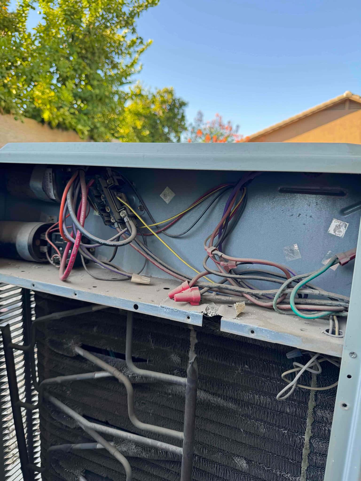 Outdoor condenser with access panel open, showing contactor, capacitor, and connected wiring above the coil, with dust accumulation.