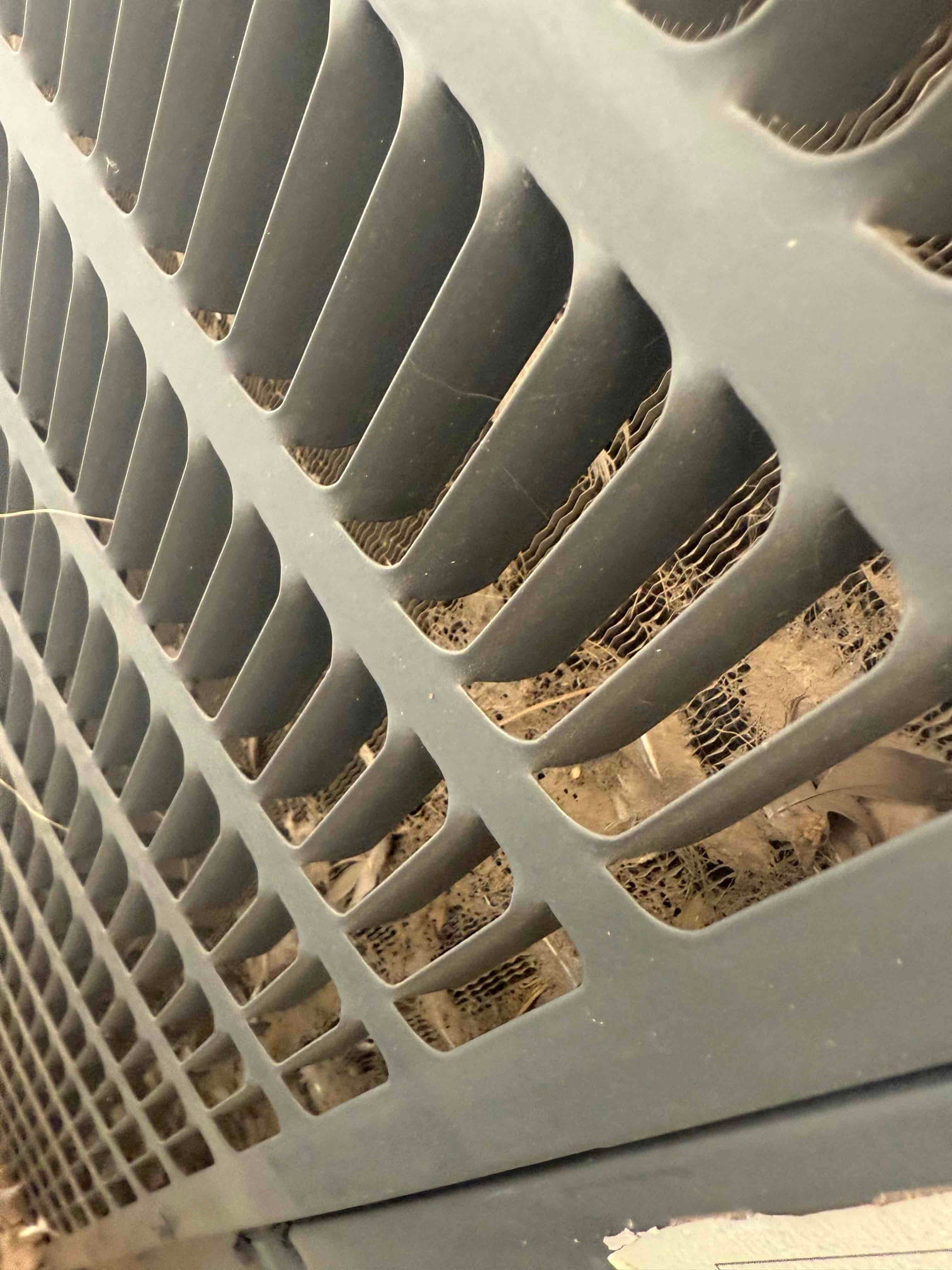 Outdoor condenser coil visible behind protective grille with accumulated lint and debris lodged in the fins.
