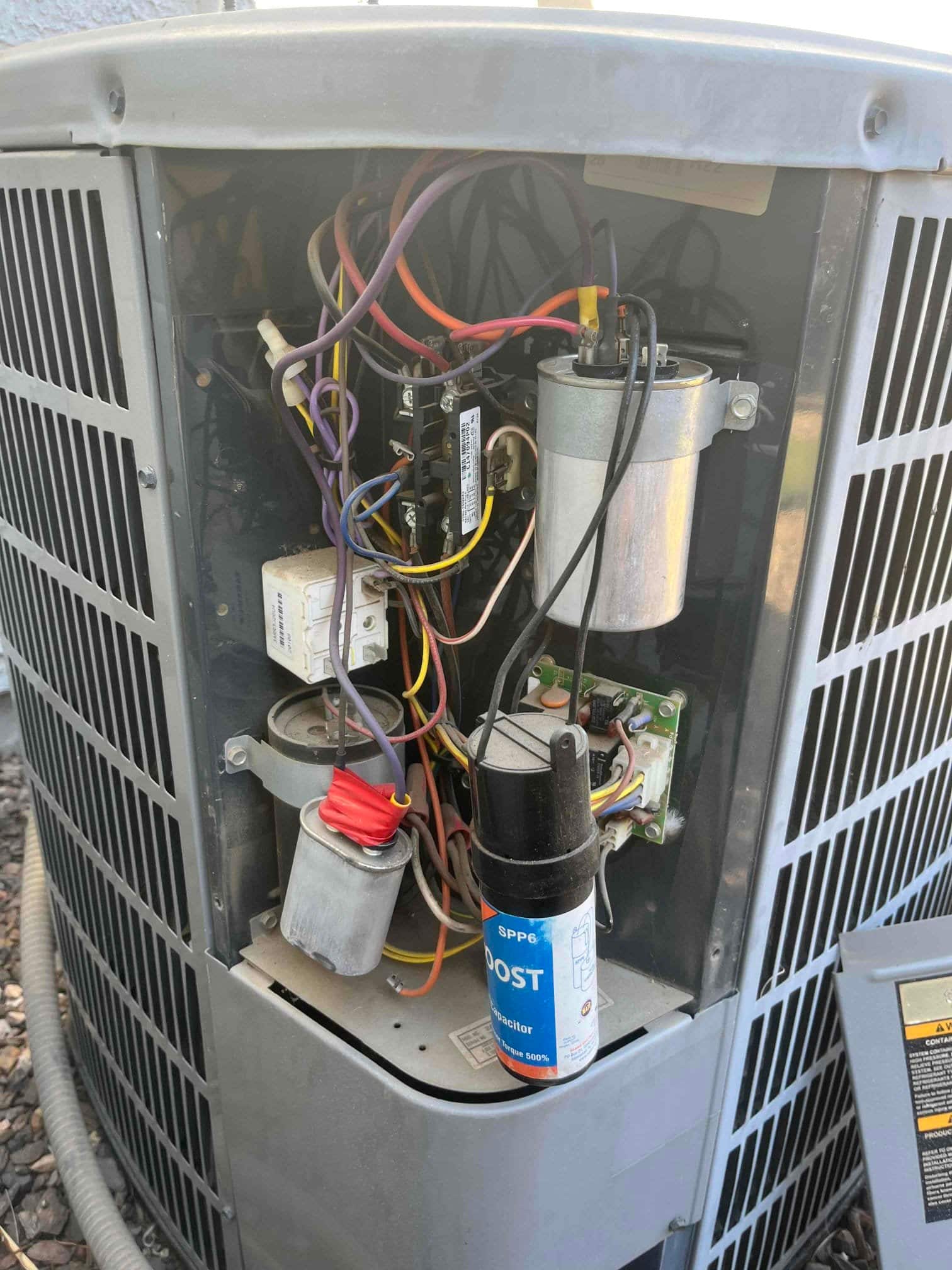 Outdoor condenser control compartment showing wiring, contactor, two cylindrical capacitors including one with red wires, and a labeled hard-start kit canister.