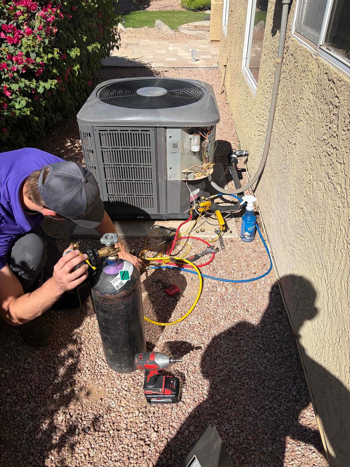 Outdoor condenser with service panel open and hoses attached, while a technician handles a cylinder beside the unit.
