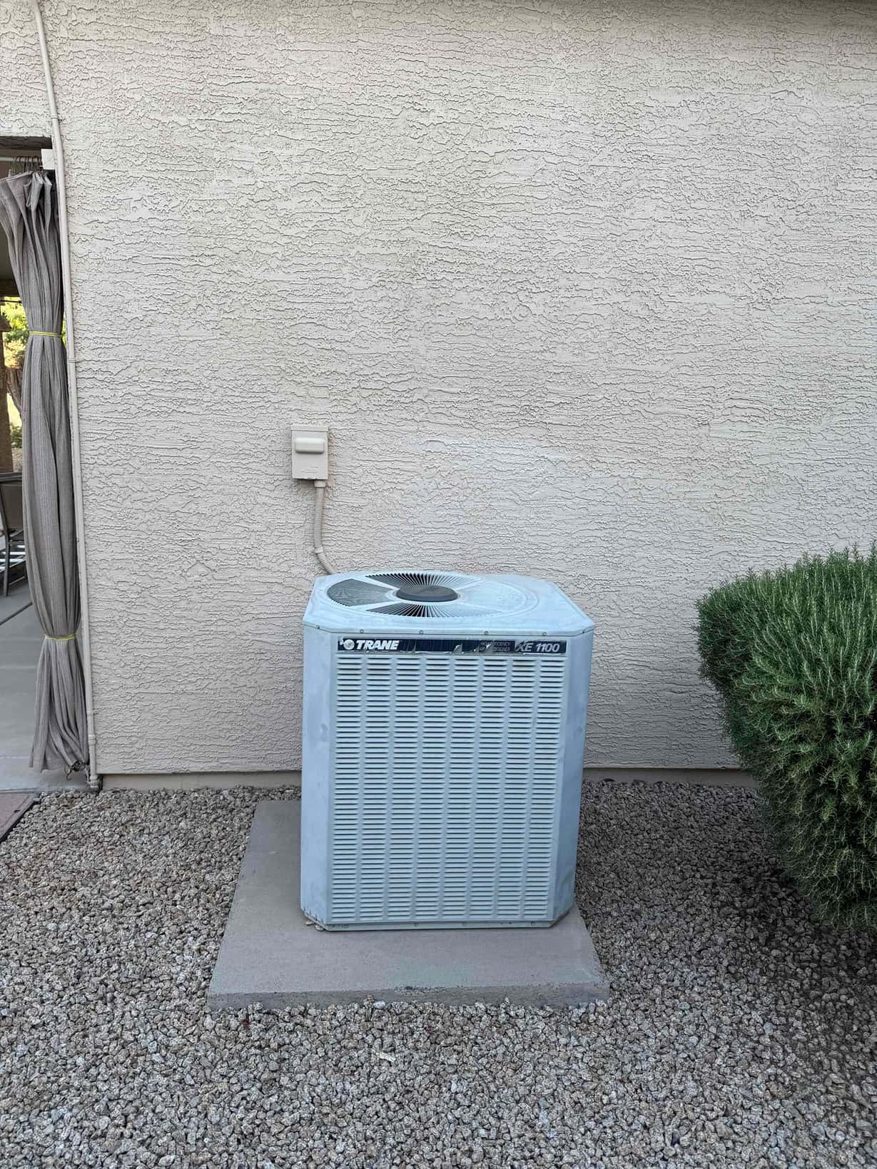 Outdoor condenser unit on concrete pad beside stucco wall with electrical whip and disconnect.
