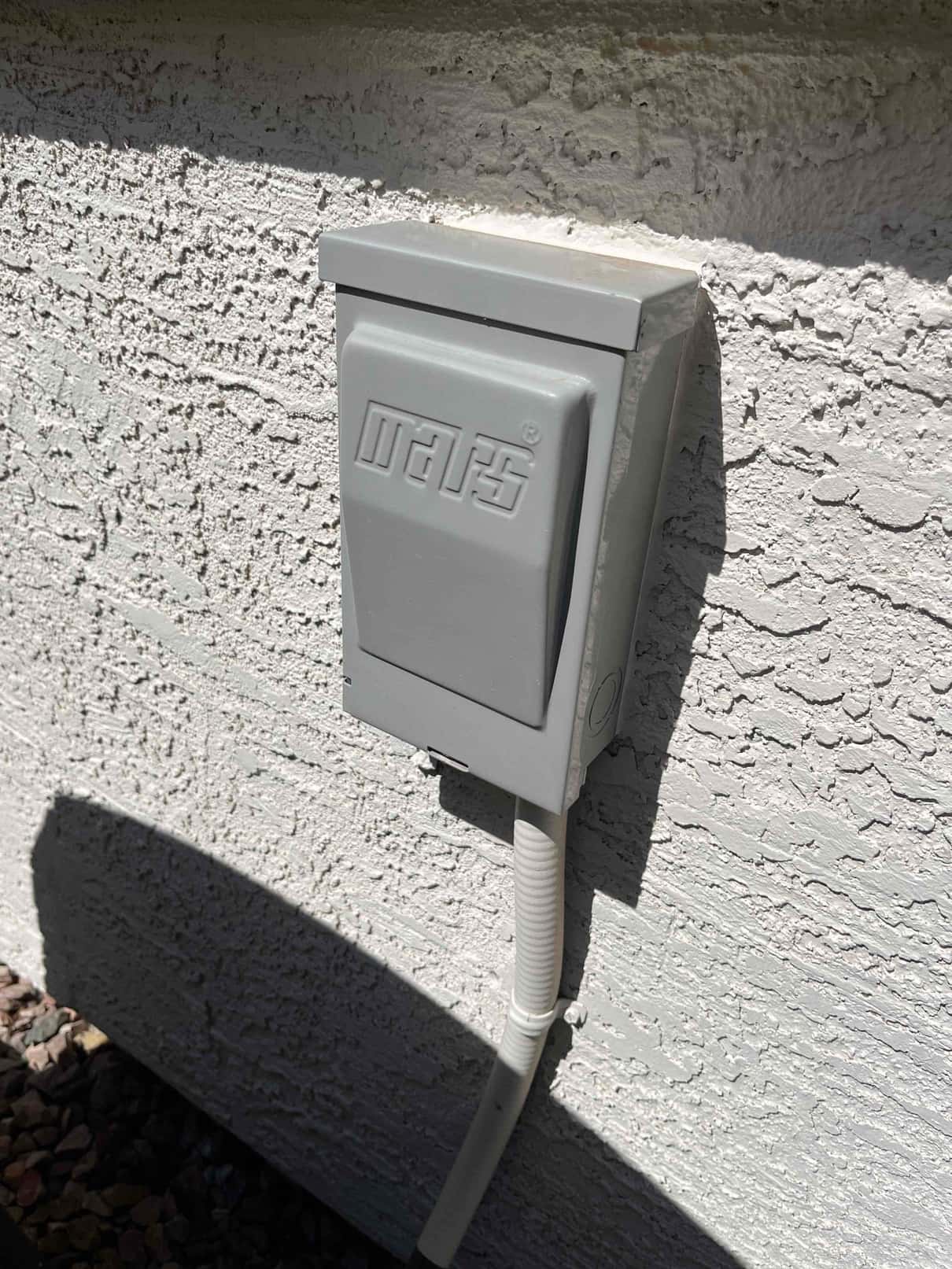 Outdoor HVAC service disconnect mounted on stucco wall with flexible conduit entering from below with cover closed.