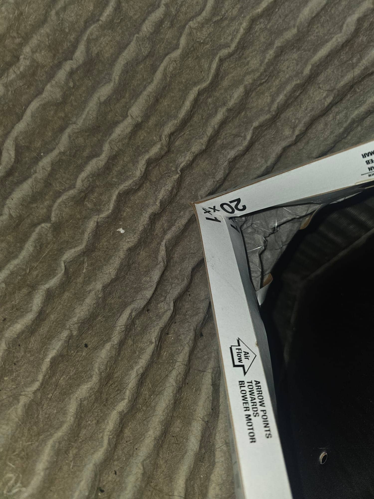 Pleated HVAC air filter with visible dust accumulation; frame shows airflow arrow pointing toward blower motor and size marking.