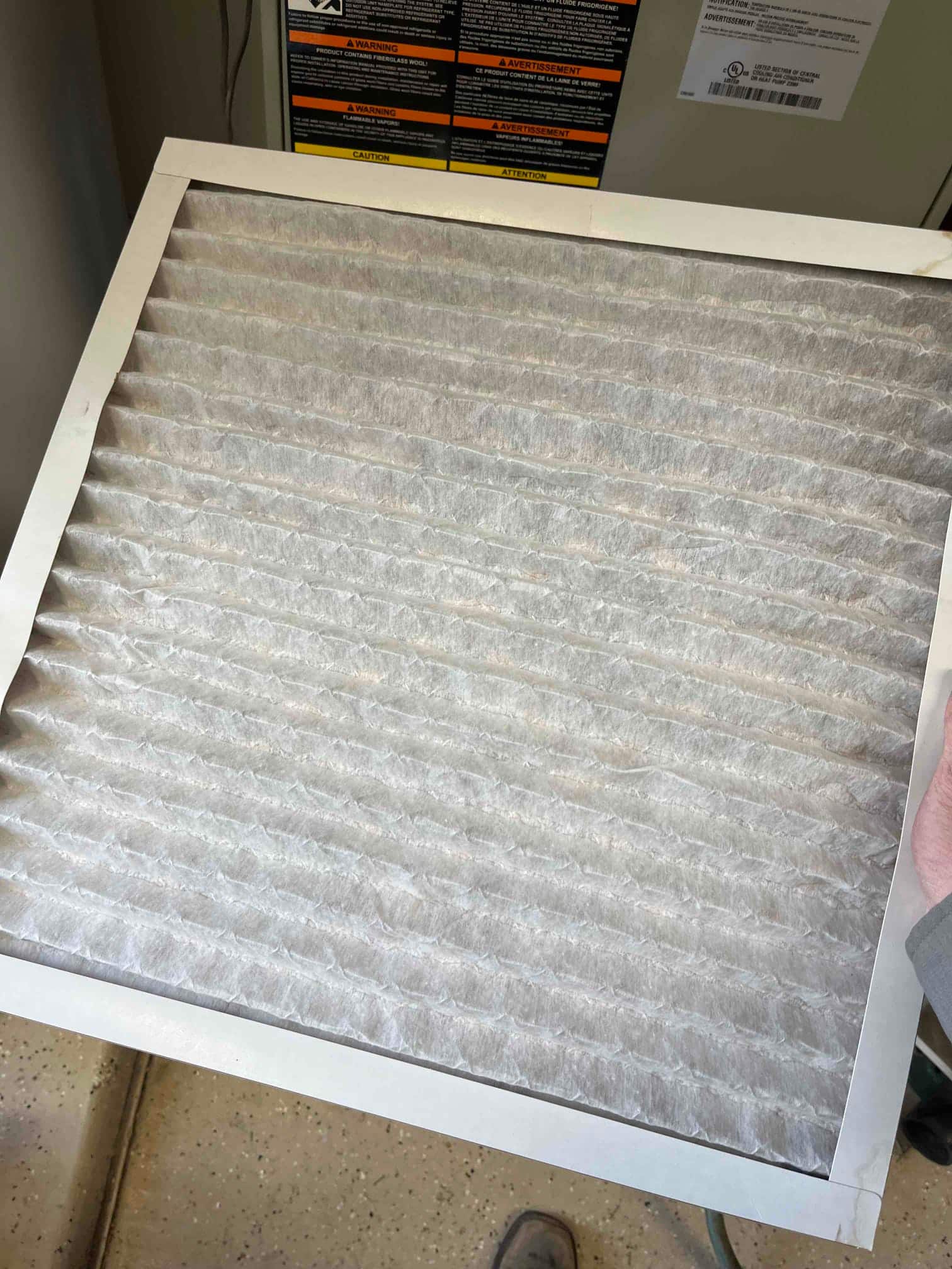 Pleated HVAC air filter held in front of an air handler cabinet with warning labels visible.