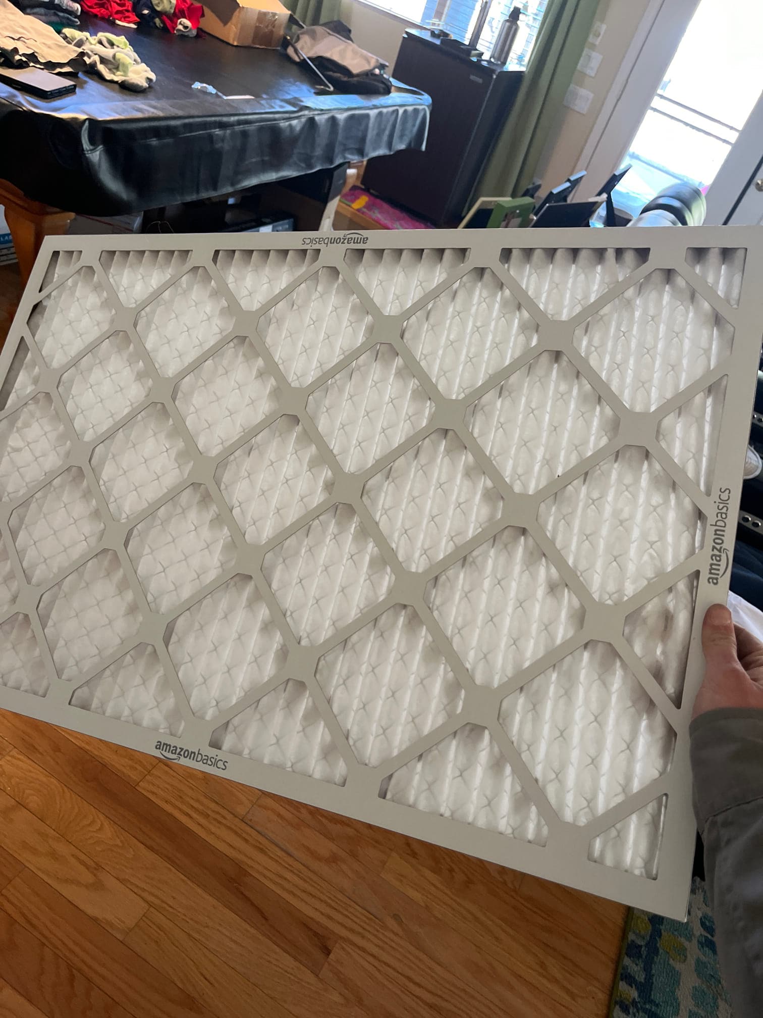 Pleated rectangular HVAC air filter with cardboard frame, held by a hand inside a residential room.