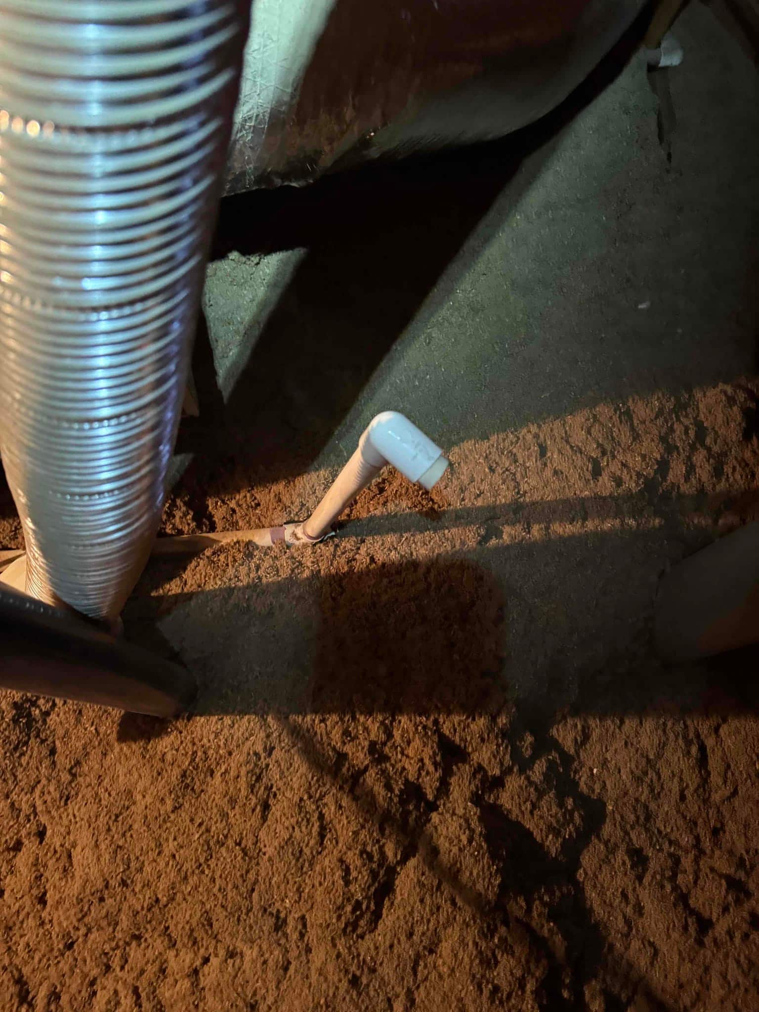 PVC condensate drain line with 90-degree elbow in crawlspace next to insulated flexible duct.