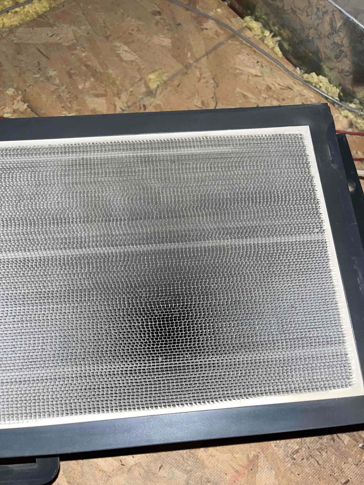 Rectangular metal mesh filter panel with plastic frame rests on attic subfloor near ductwork.