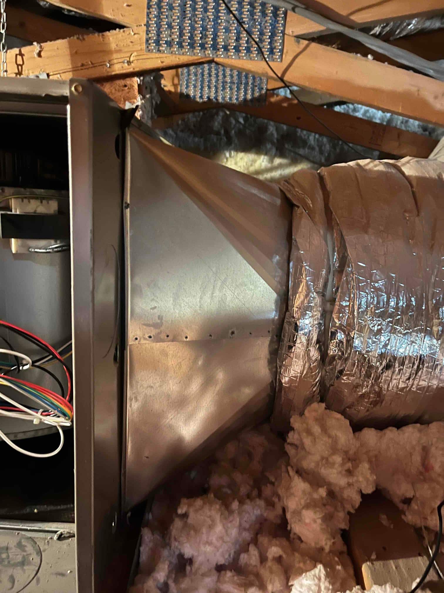 Sheet-metal transition duct connected to insulated flexible duct at attic air handler, with exposed fiberglass insulation.