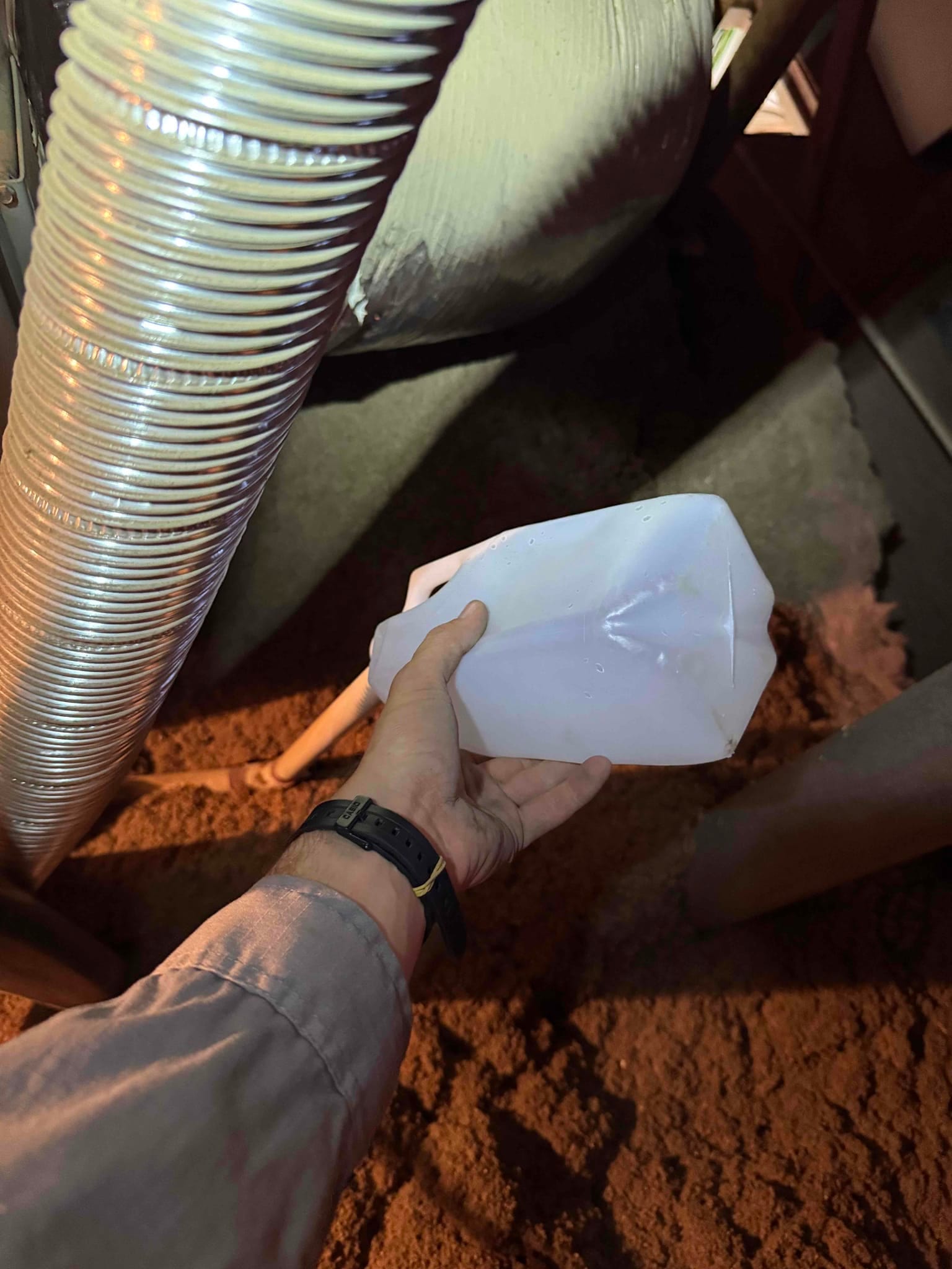 Technician holds a container beneath a small PVC drain line near insulated ductwork in a crawlspace.