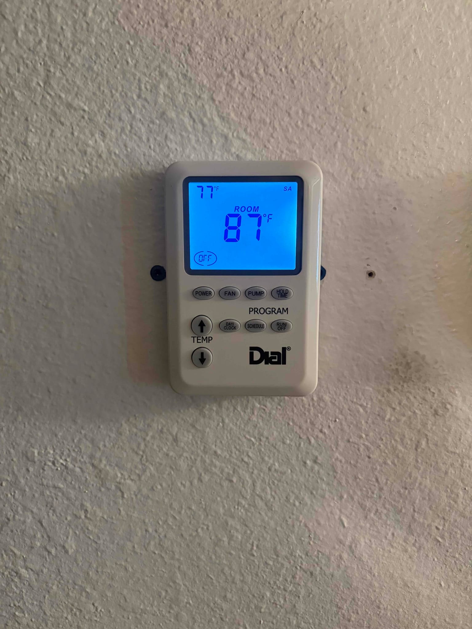 Wall-mounted thermostat with backlit display reading ROOM 87°F, with 77°F shown smaller at the upper left.