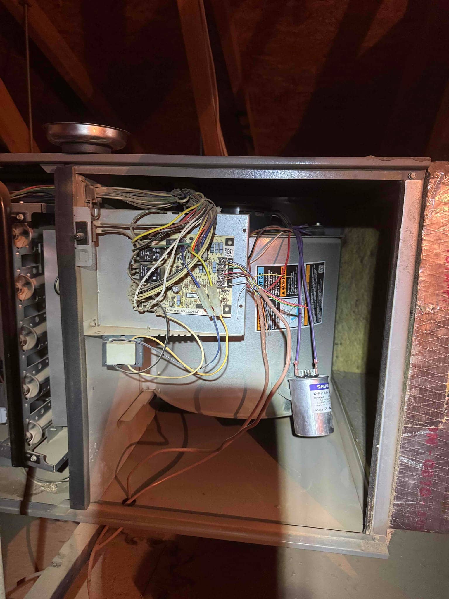 Furnace cabinet open in attic, showing control board, wiring bundle, blower housing, and a mounted run capacitor.