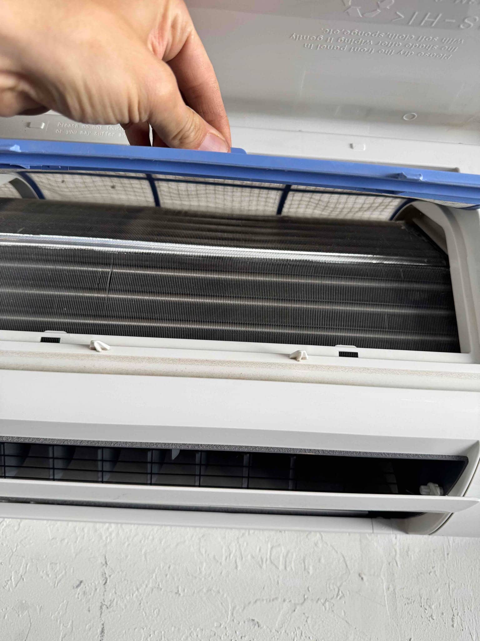 A hand lifts the blue filter frame on a wall-mounted indoor unit, exposing evaporator coil.