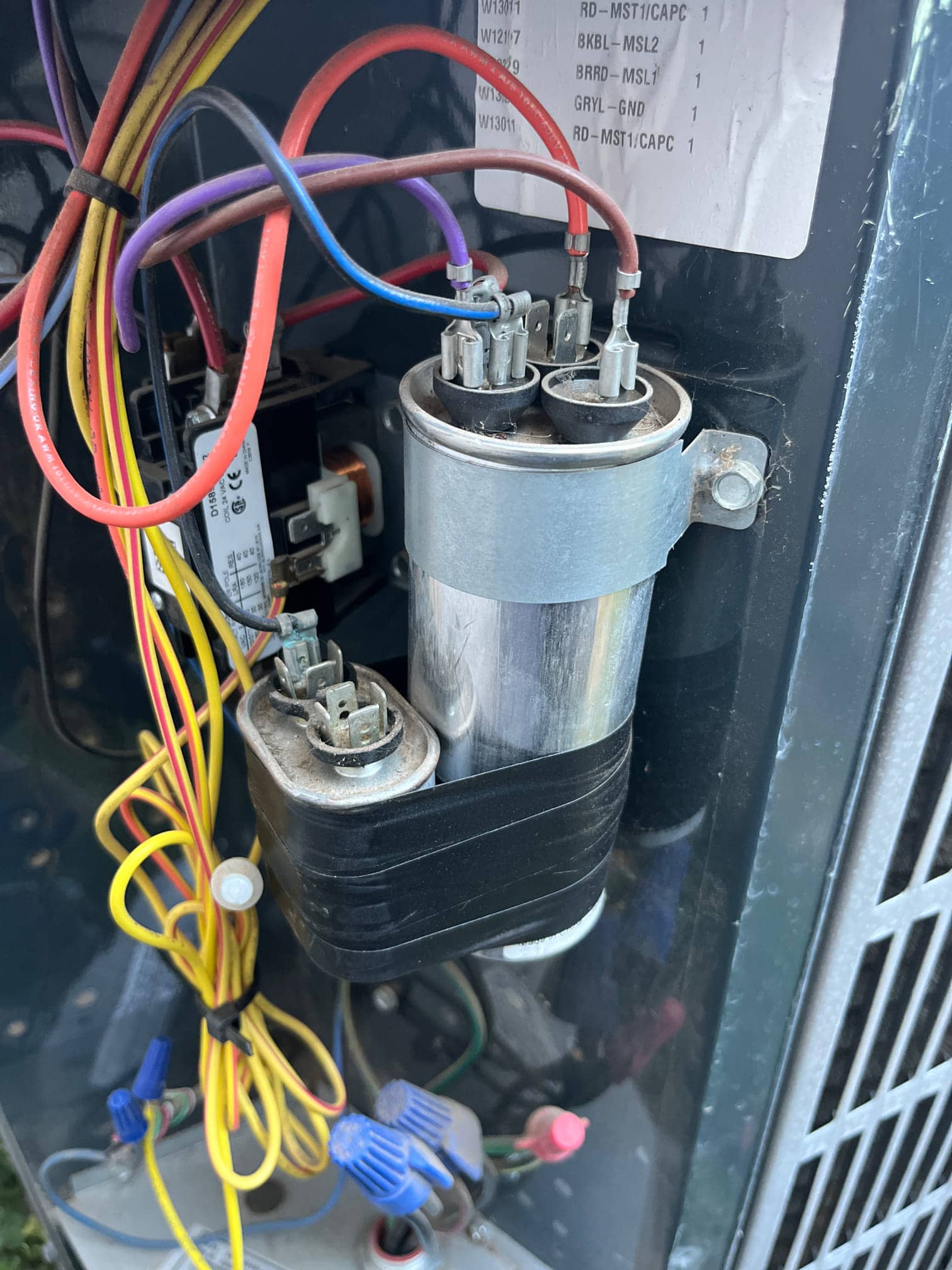 Two capacitors are mounted in the outdoor condenser compartment, with connected wires, nearby contactor, and bundled yellow leads.